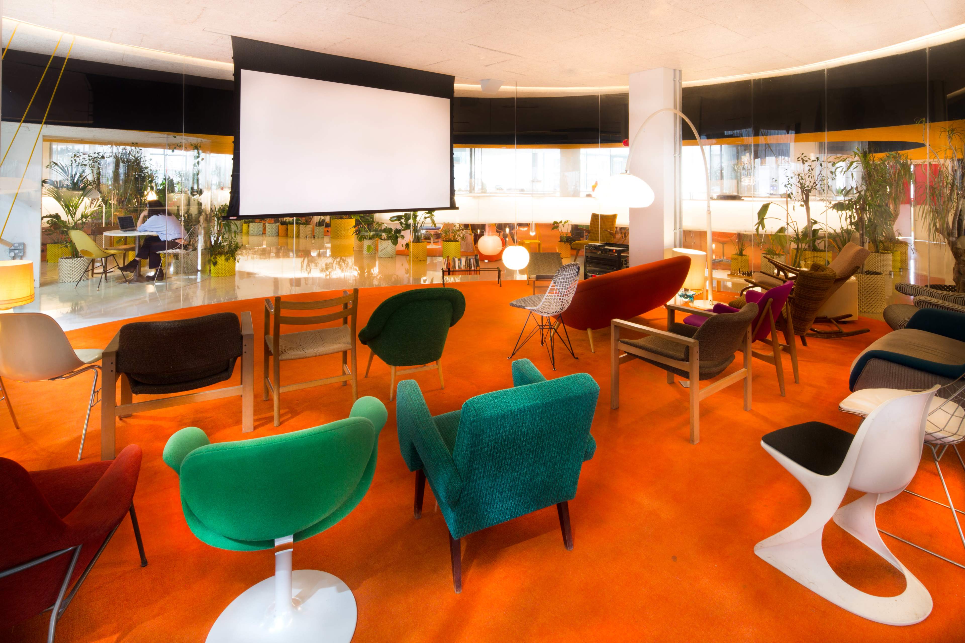 A spacious lounge area features a variety of colorful chairs arranged facing a large projection screen, with plants and modern decor visible in the background.