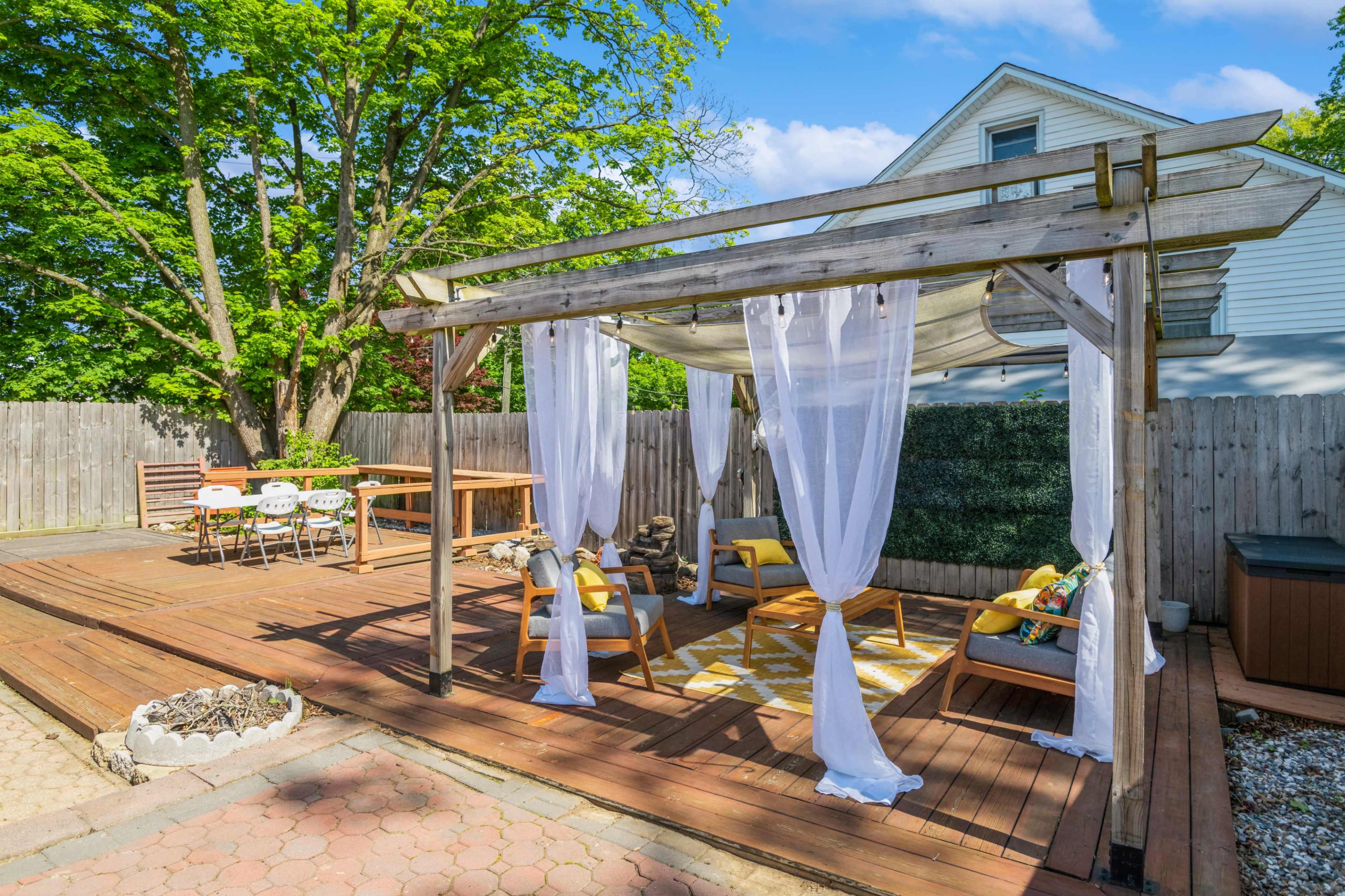 The image shows a wooden patio structure with white drapes, seating areas, and a dining table surrounded by greenery and a wooden fence.