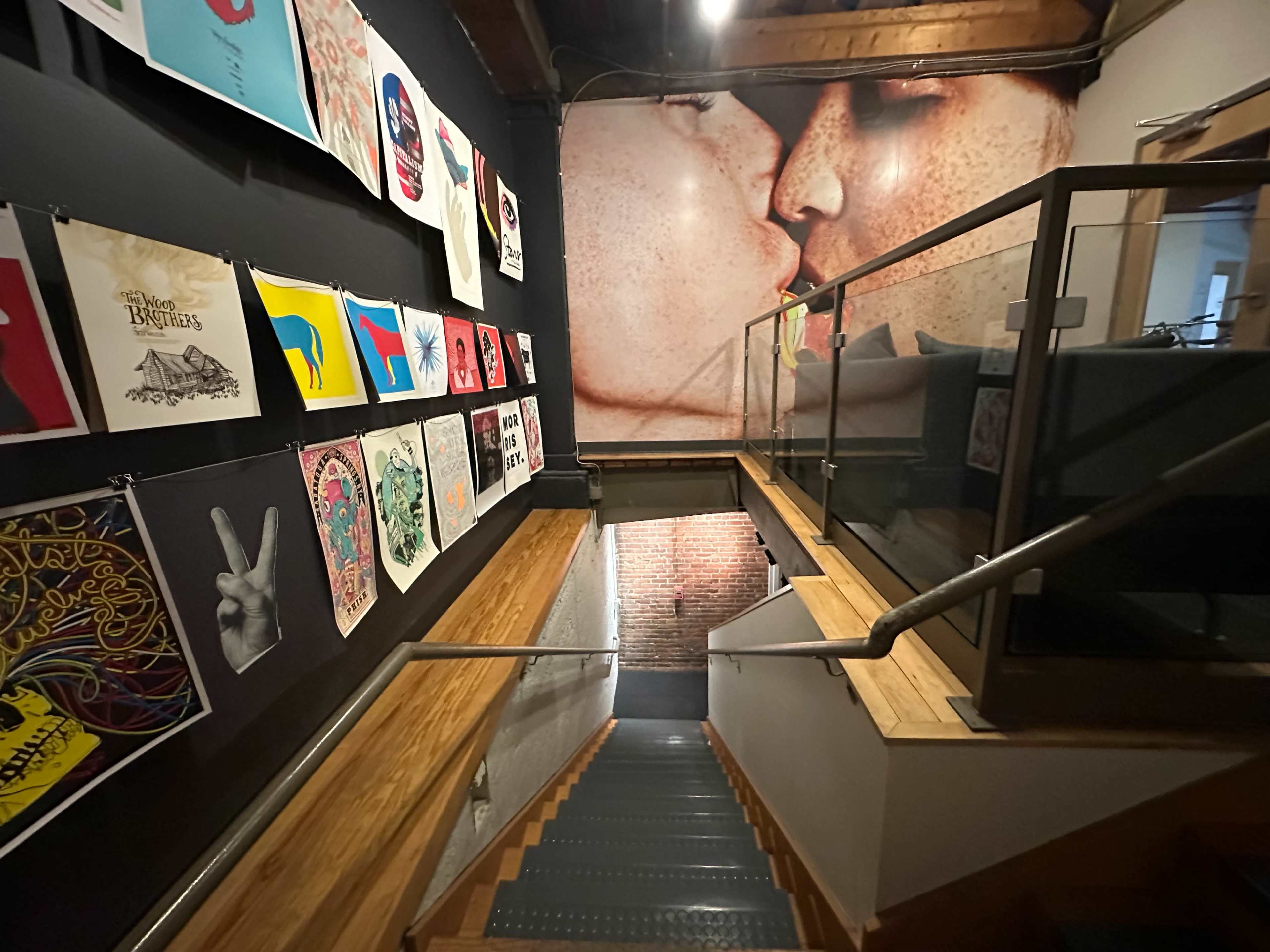 A stairway leading down with wooden steps, flanked by artwork on the walls, and a large mural of a kiss at the end of the corridor.