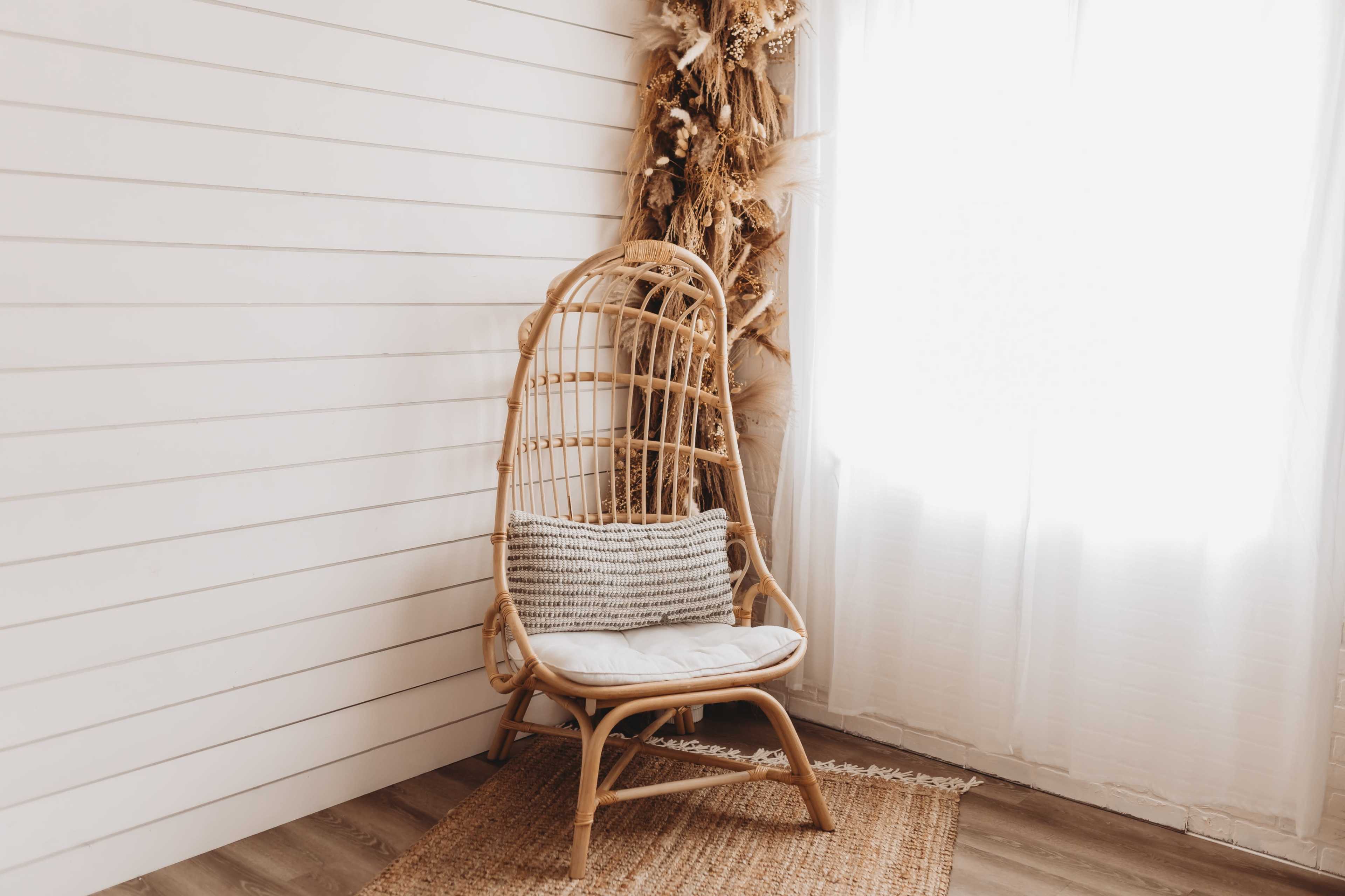 A rattan chair is positioned in a bright room with white wood paneling and a light curtain, surrounded by decorative dried plants.