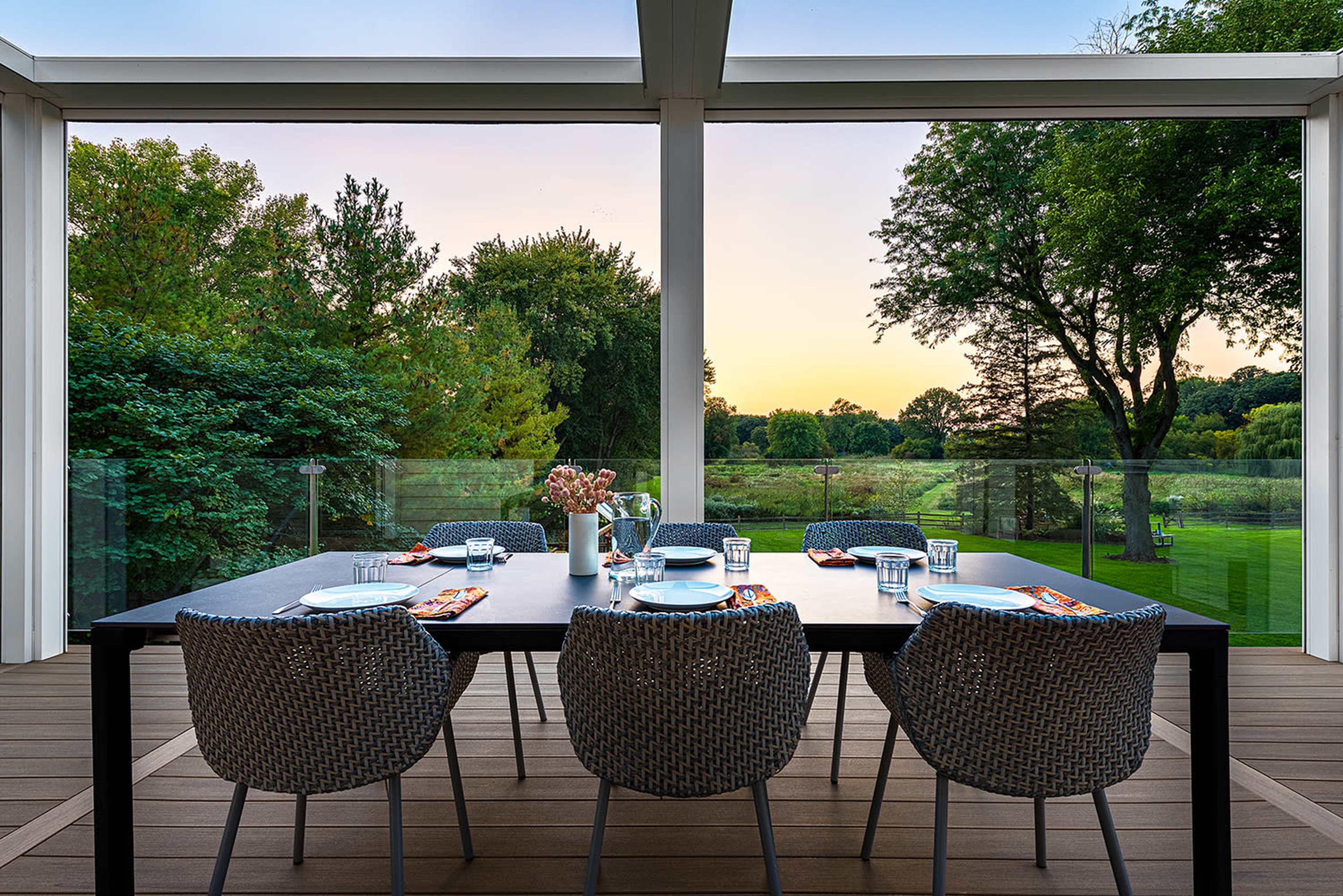 A modern dining table is set on a patio, overlooking a green landscape at sunset, with chairs arranged around it.