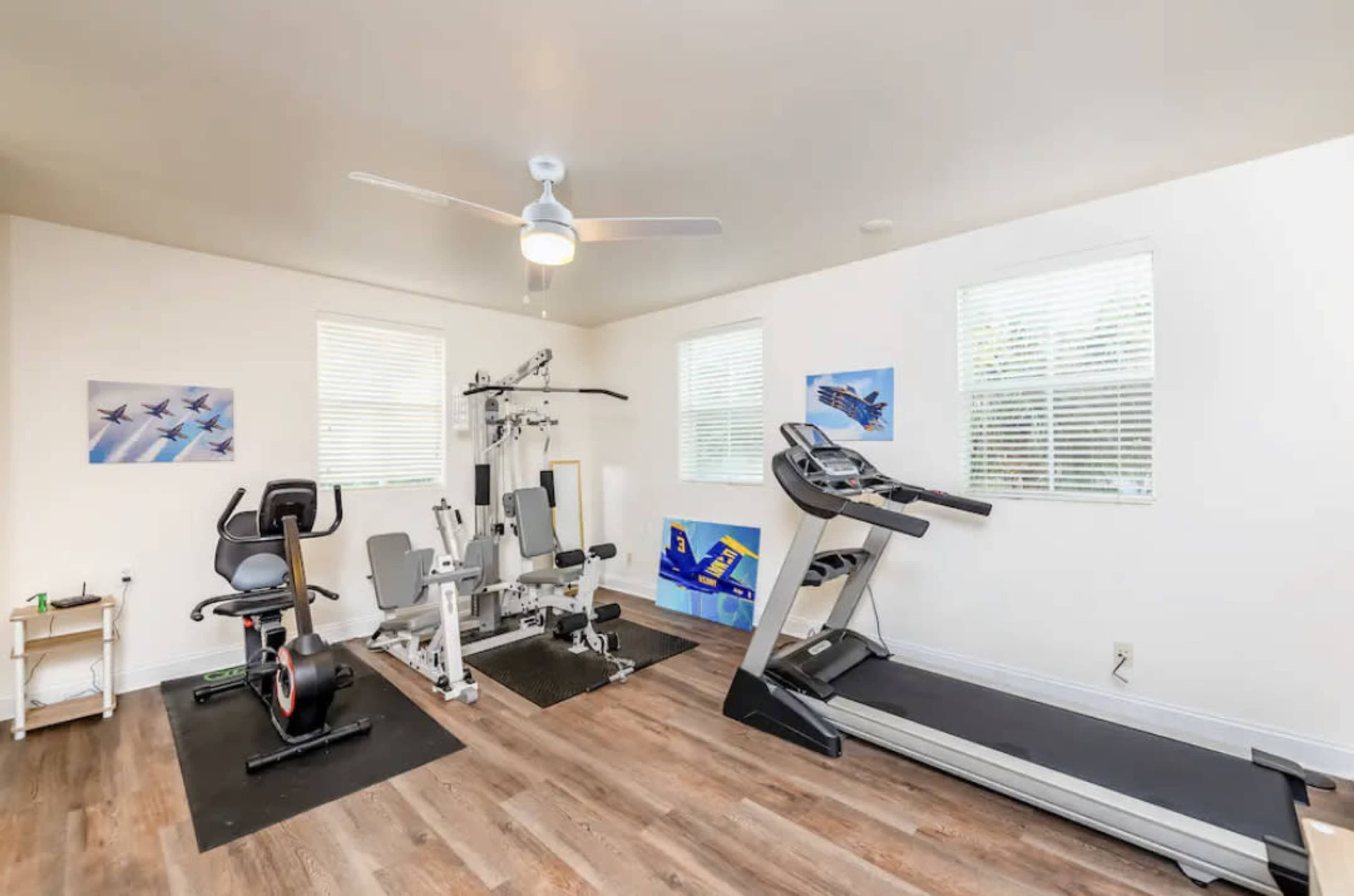 A home gym with exercise equipment including a multi-station gym machine, a stationary bike, and a treadmill, set in a well-lit room with windows and artwork on the walls.