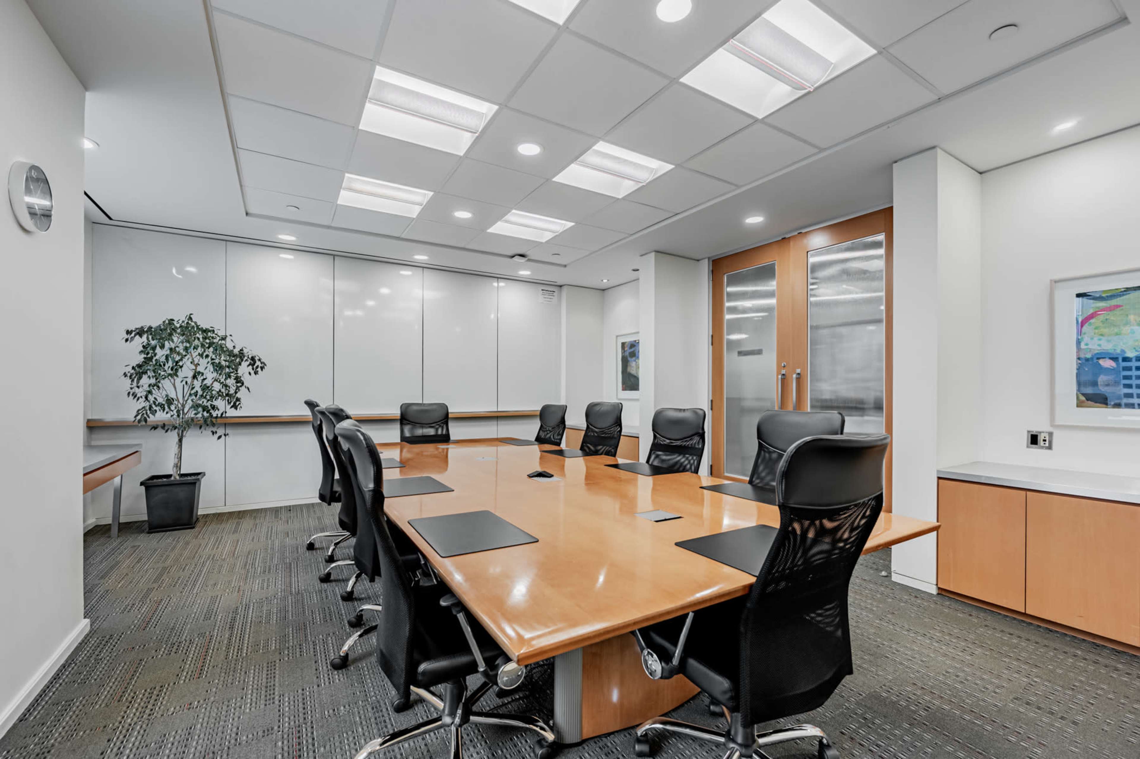 A modern conference room features a long wooden table surrounded by black ergonomic chairs, with a large whiteboard and glass doors in the background.