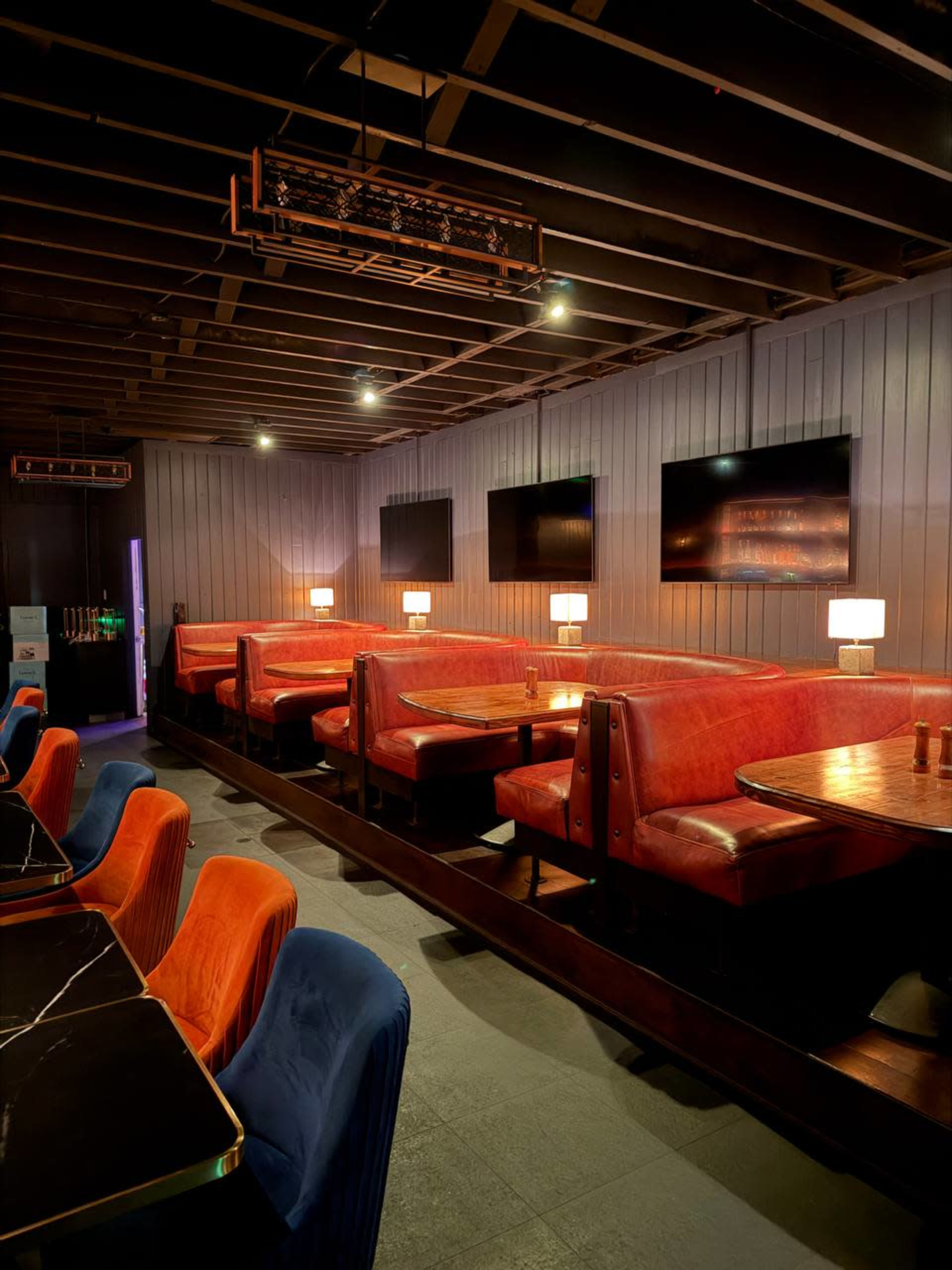 The image shows a modern restaurant interior with red and blue seating booths, wooden tables, and several wall-mounted televisions.