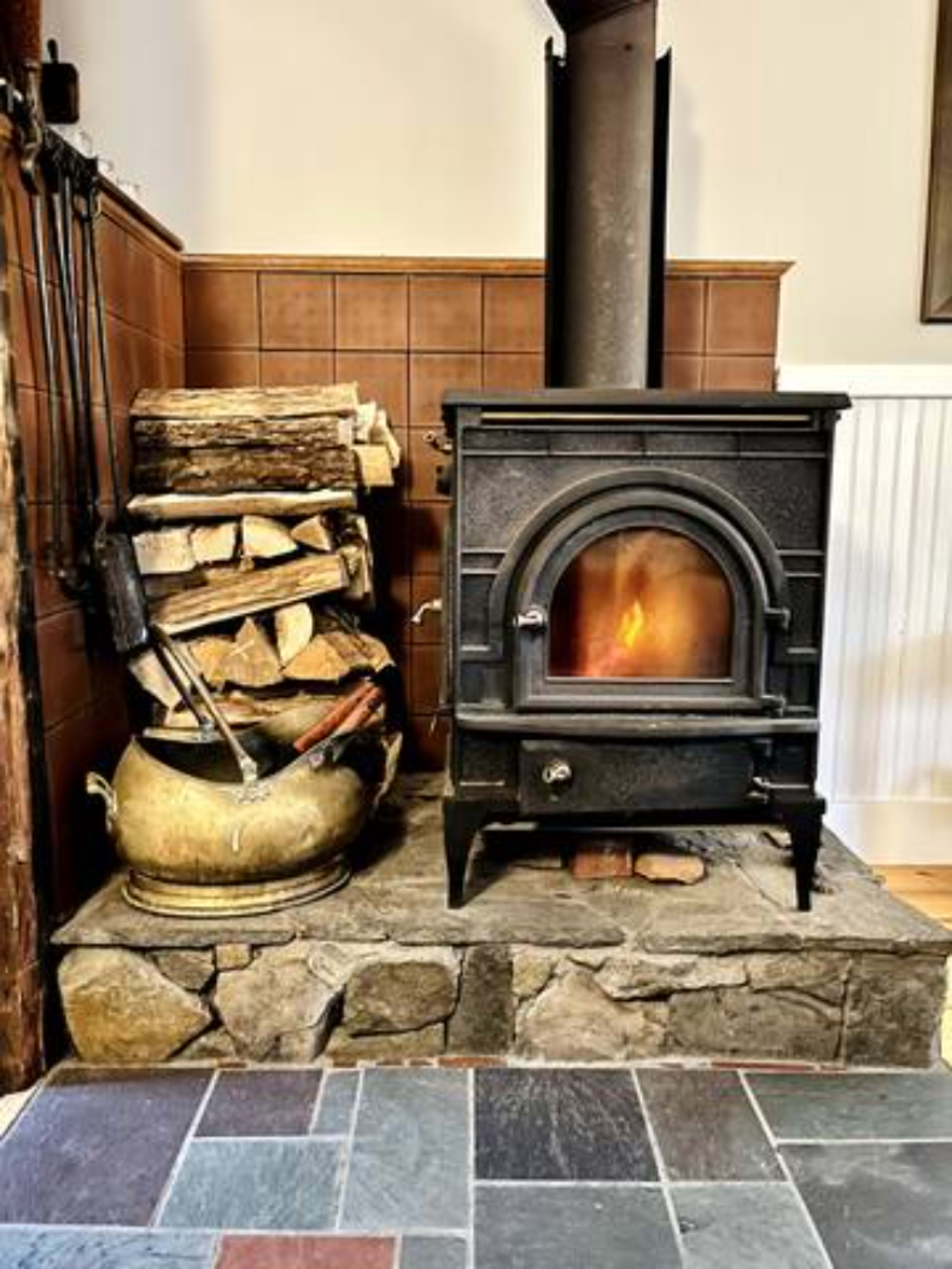A rustic wood stove beside a stacked pile of firewood and an antique pot on a stone base.