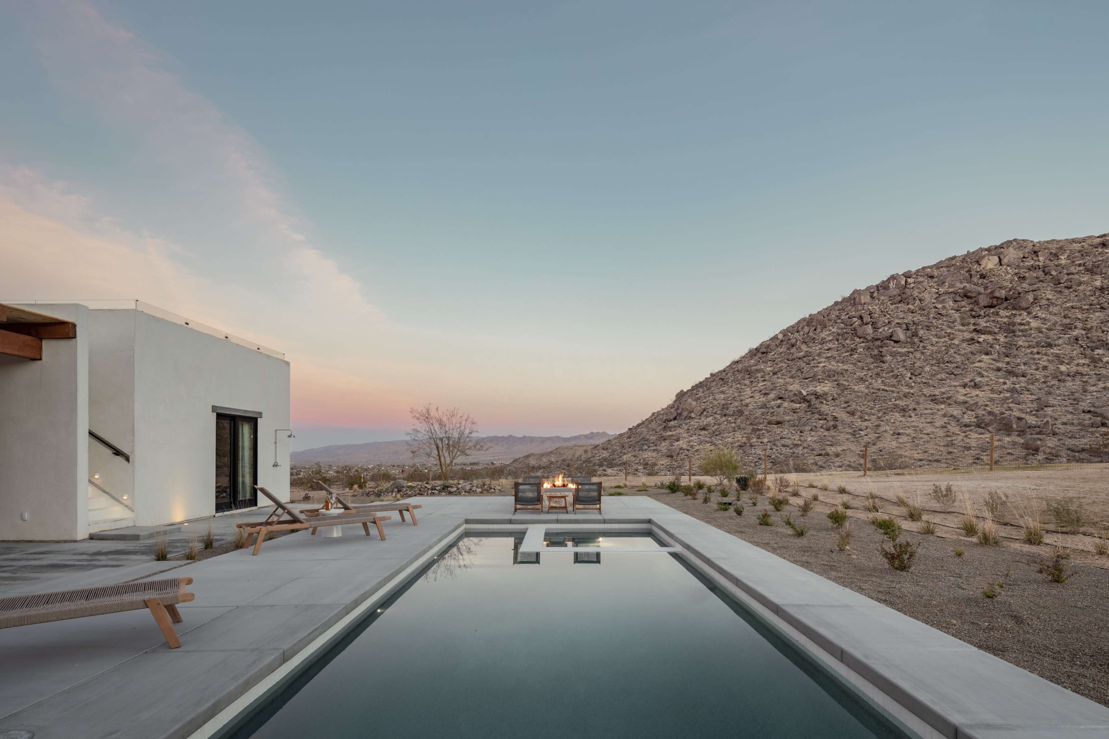 A modern home with a pool is situated near a rocky hillside, surrounded by sparse desert landscaping under a pastel sky.
