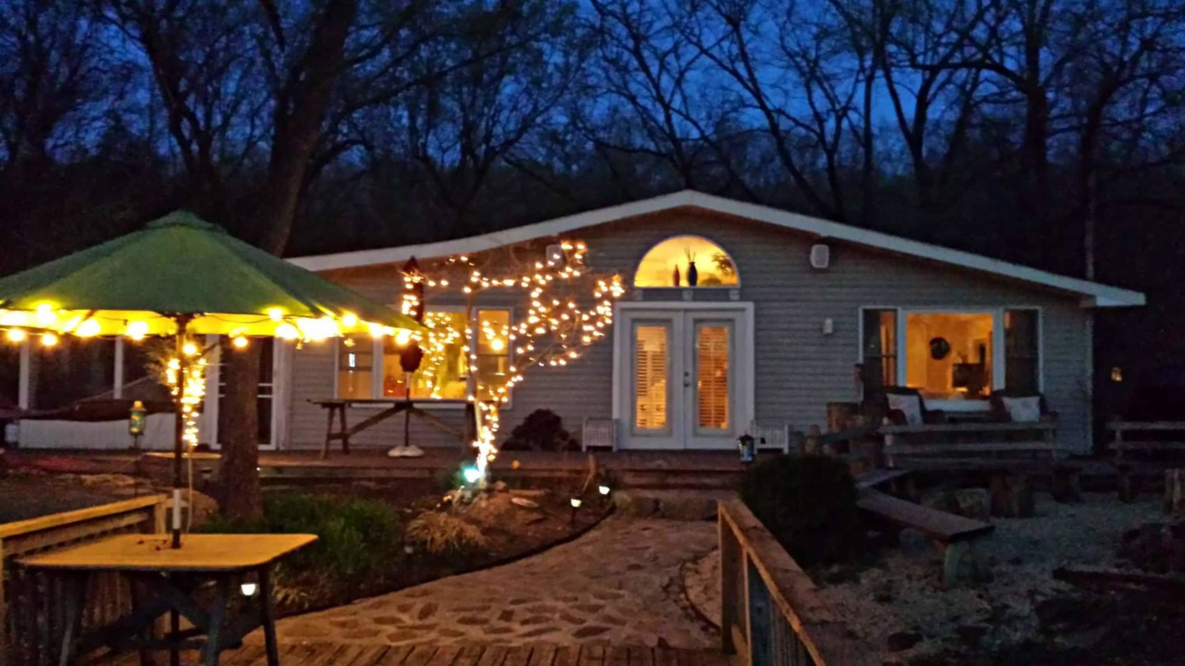 A cozy house with string lights wrapped around a tree is nestled in a wooded area at dusk, with a green umbrella-covered table in the foreground.