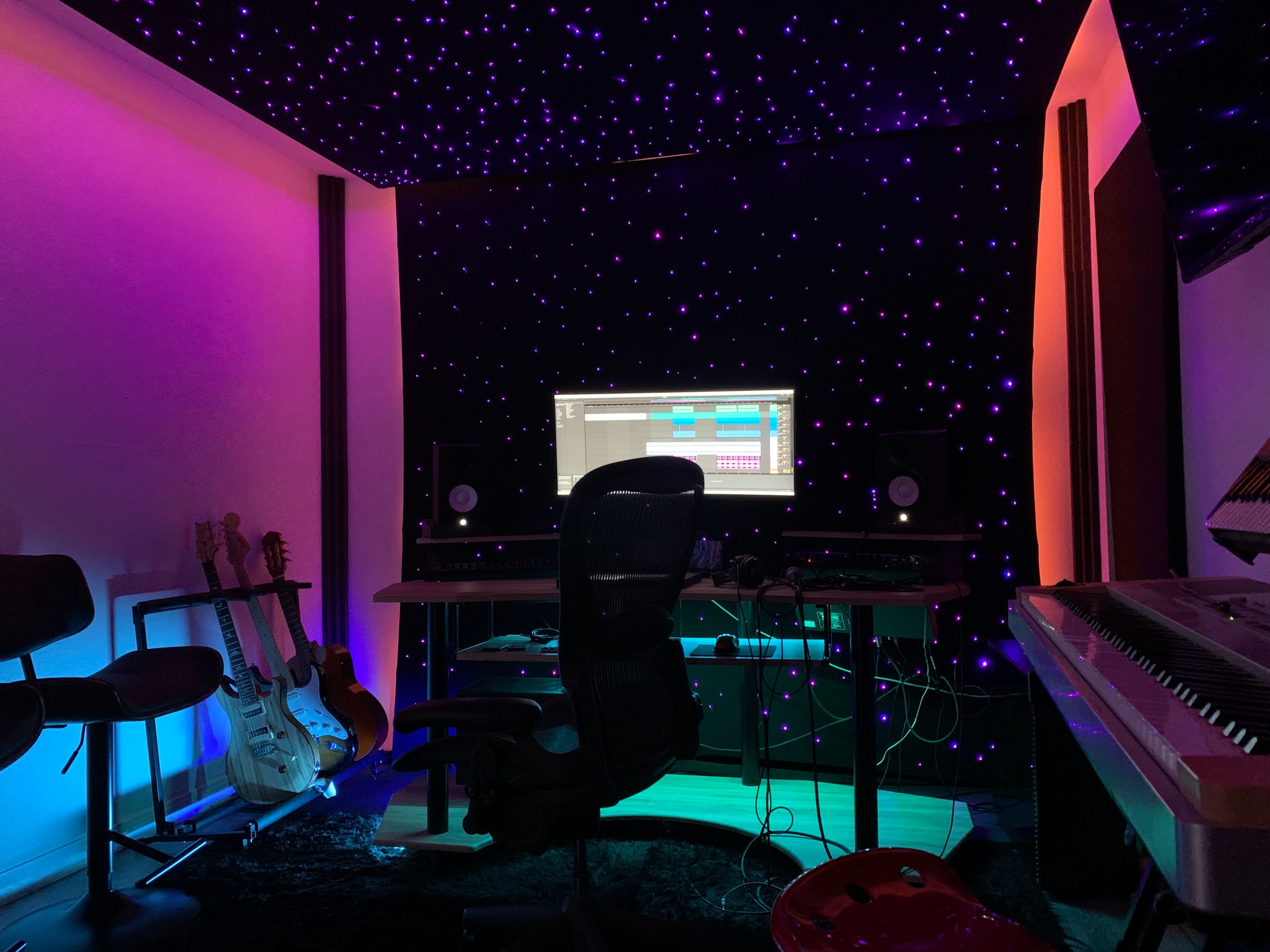 A music studio features a computer setup with monitors, surrounded by guitars and illuminated by colorful lights against a starry backdrop.