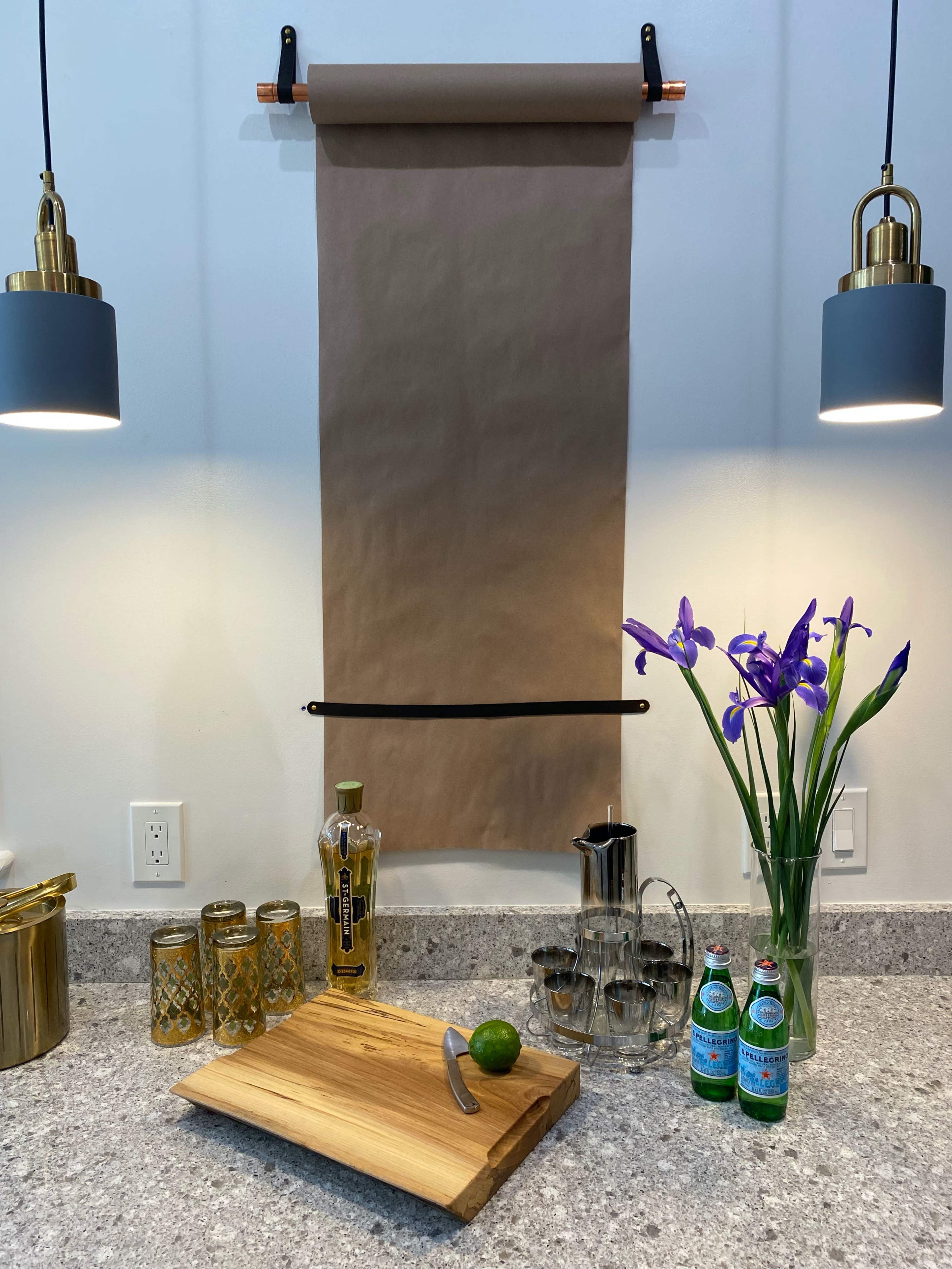 A wooden cutting board with a lime and a knife sits on a countertop below a hanging sheet of brown paper, accompanied by a pitcher, glassware, and two bottles of sparkling water alongside a vase of purple flowers.