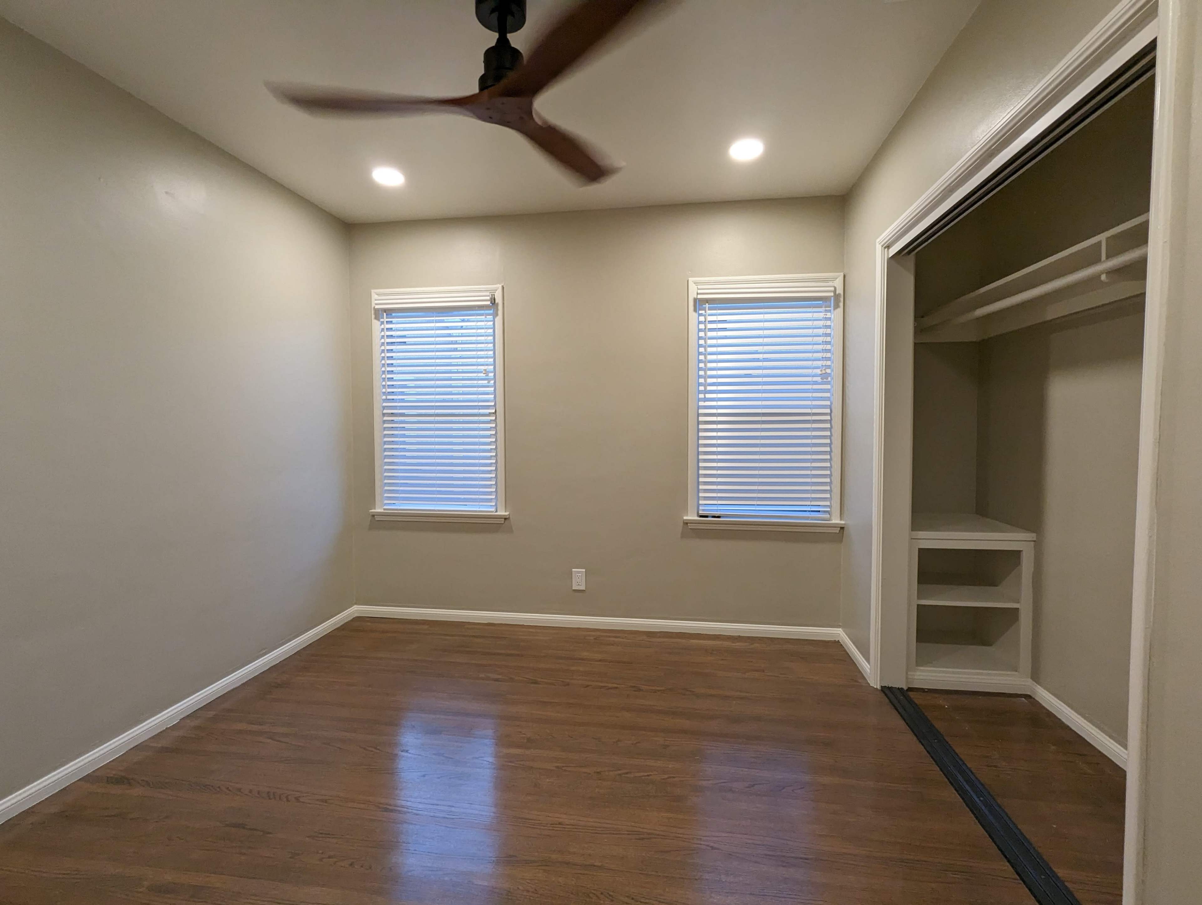 The image shows a small, empty room with hardwood flooring, two windows with blinds, and a closet with open shelving.
