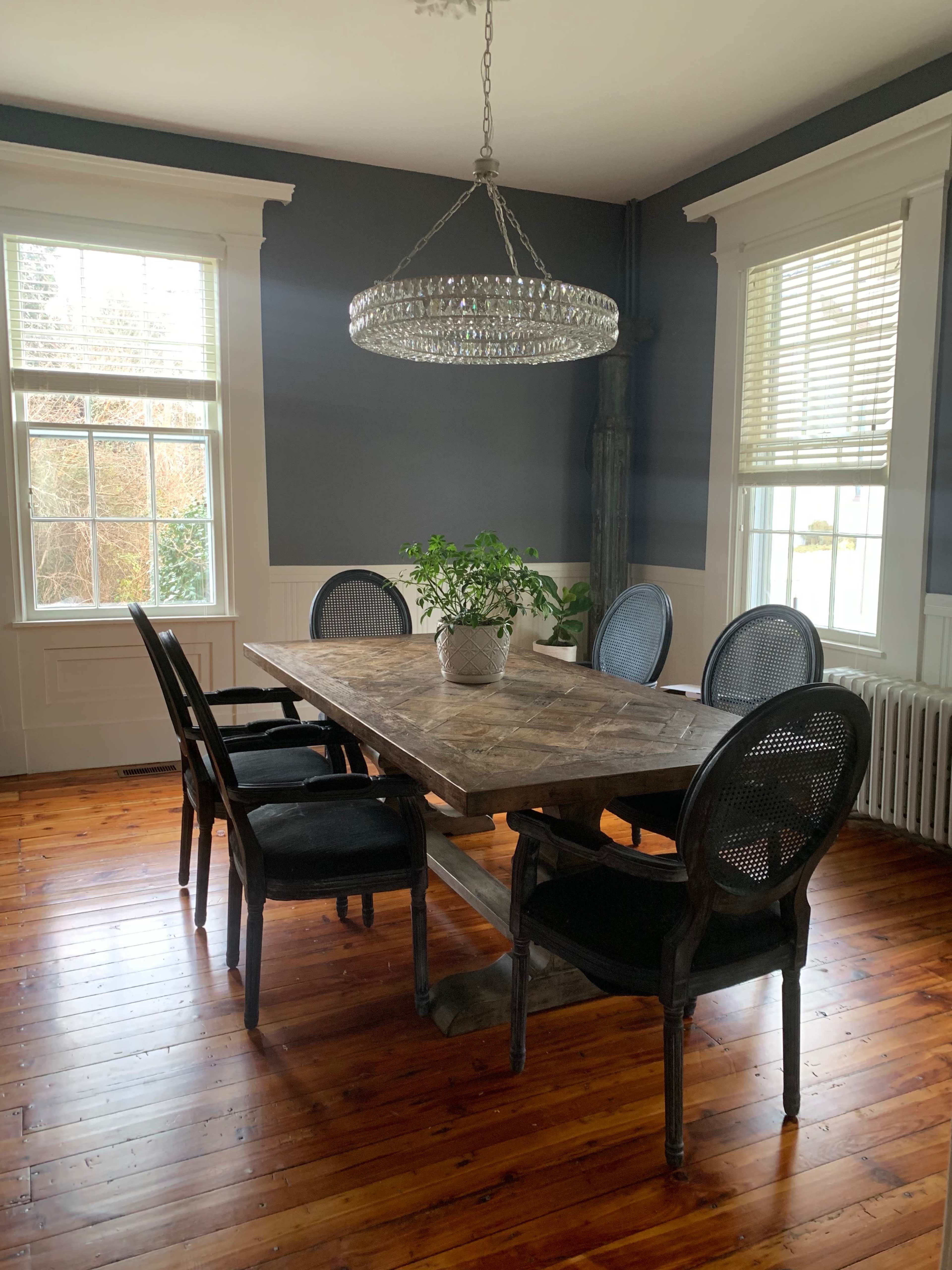 A wooden dining table with six chairs and a large chandelier is positioned in a well-lit room with blue walls and wooden flooring.