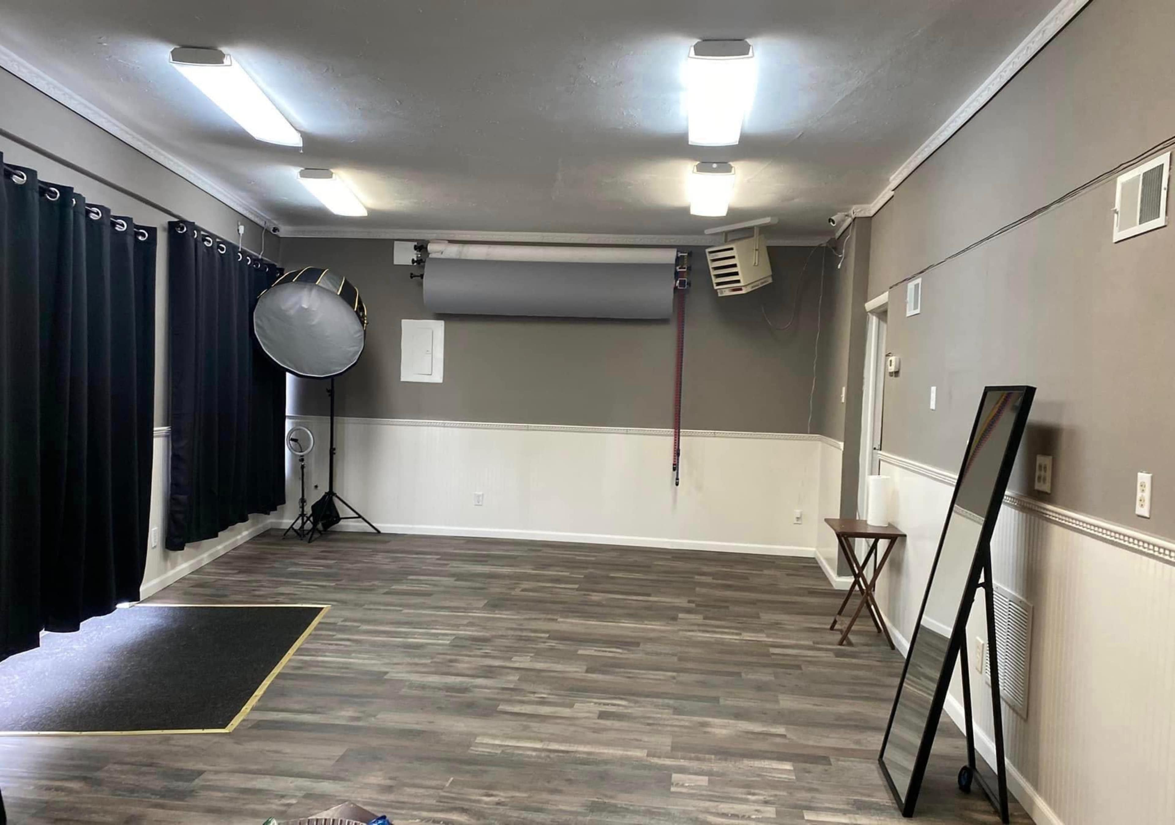 A spacious, empty studio with gray walls, wooden flooring, a large reflector, and black curtains on the windows.