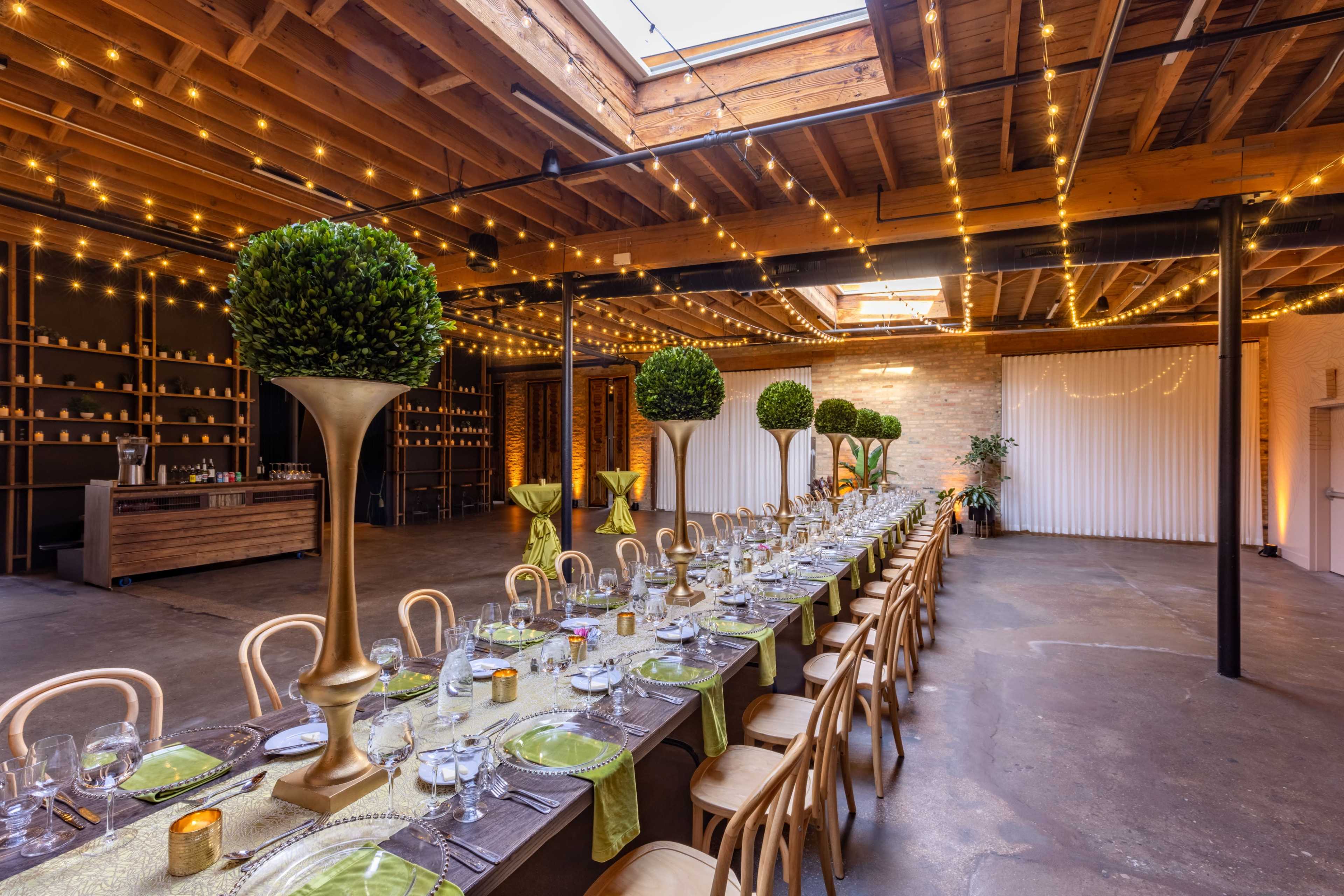 A long dining table is set with green tableware and surrounded by tall topiary arrangements in a brightly lit event space with exposed wooden beams and string lights.
