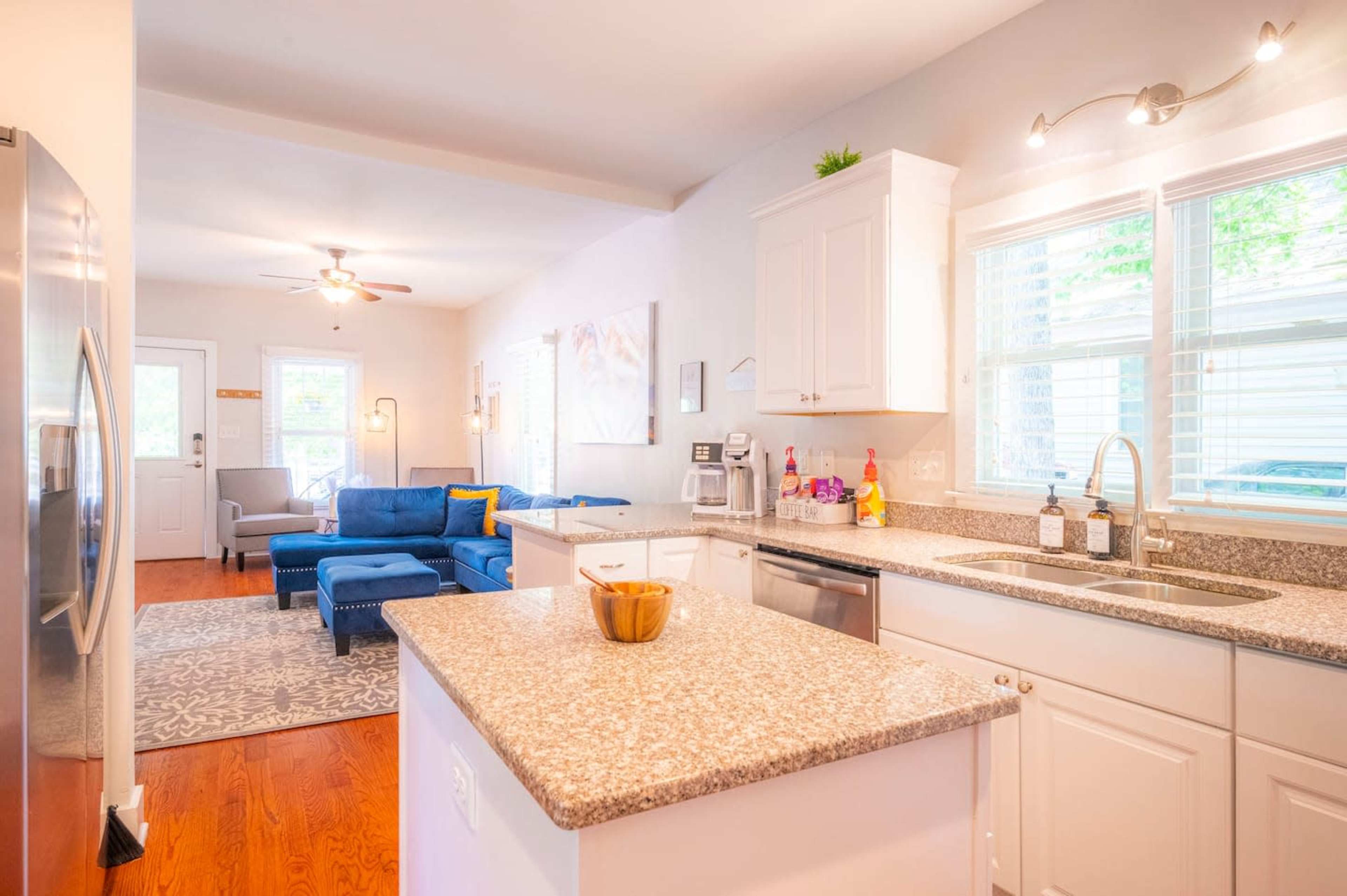 A modern kitchen with a central island and gray granite countertops opens into a cozy living area featuring a blue couch.