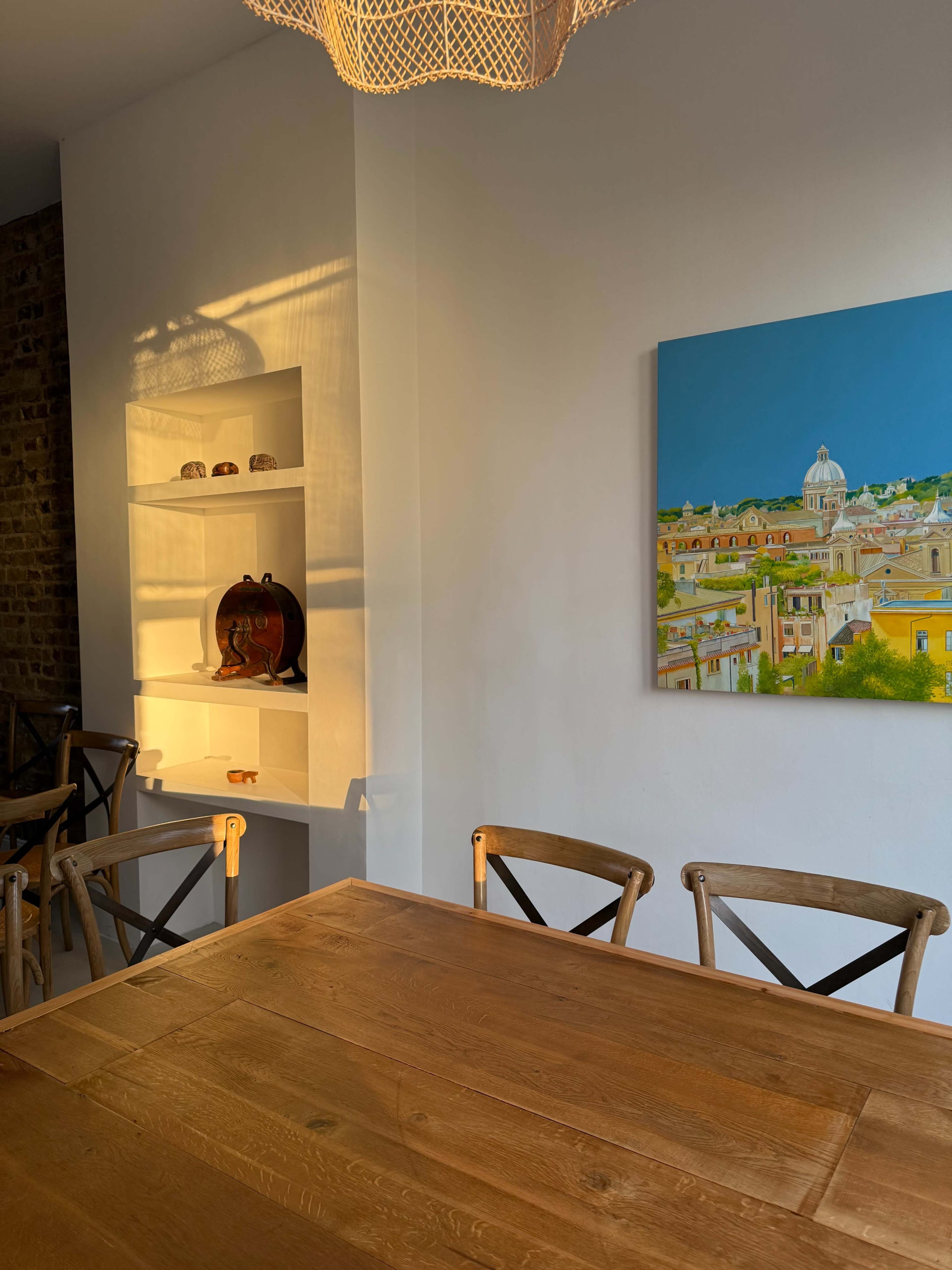 A wooden dining table with cross-back chairs is illuminated by sunlight, alongside a wall featuring a painting and a white shelf displaying decorative items.