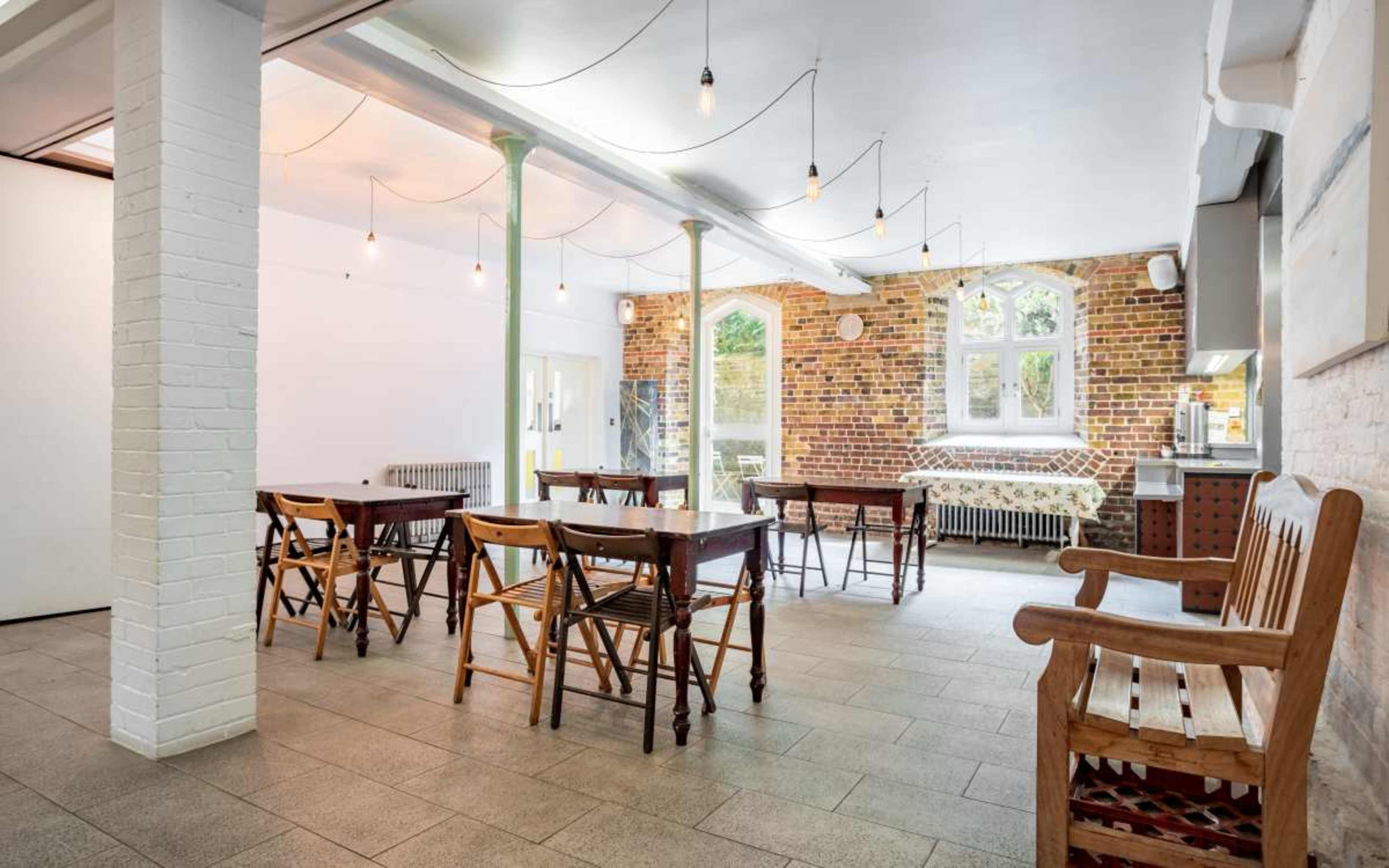 A spacious room with wooden tables and chairs, exposed brick walls, and large windows letting in natural light.