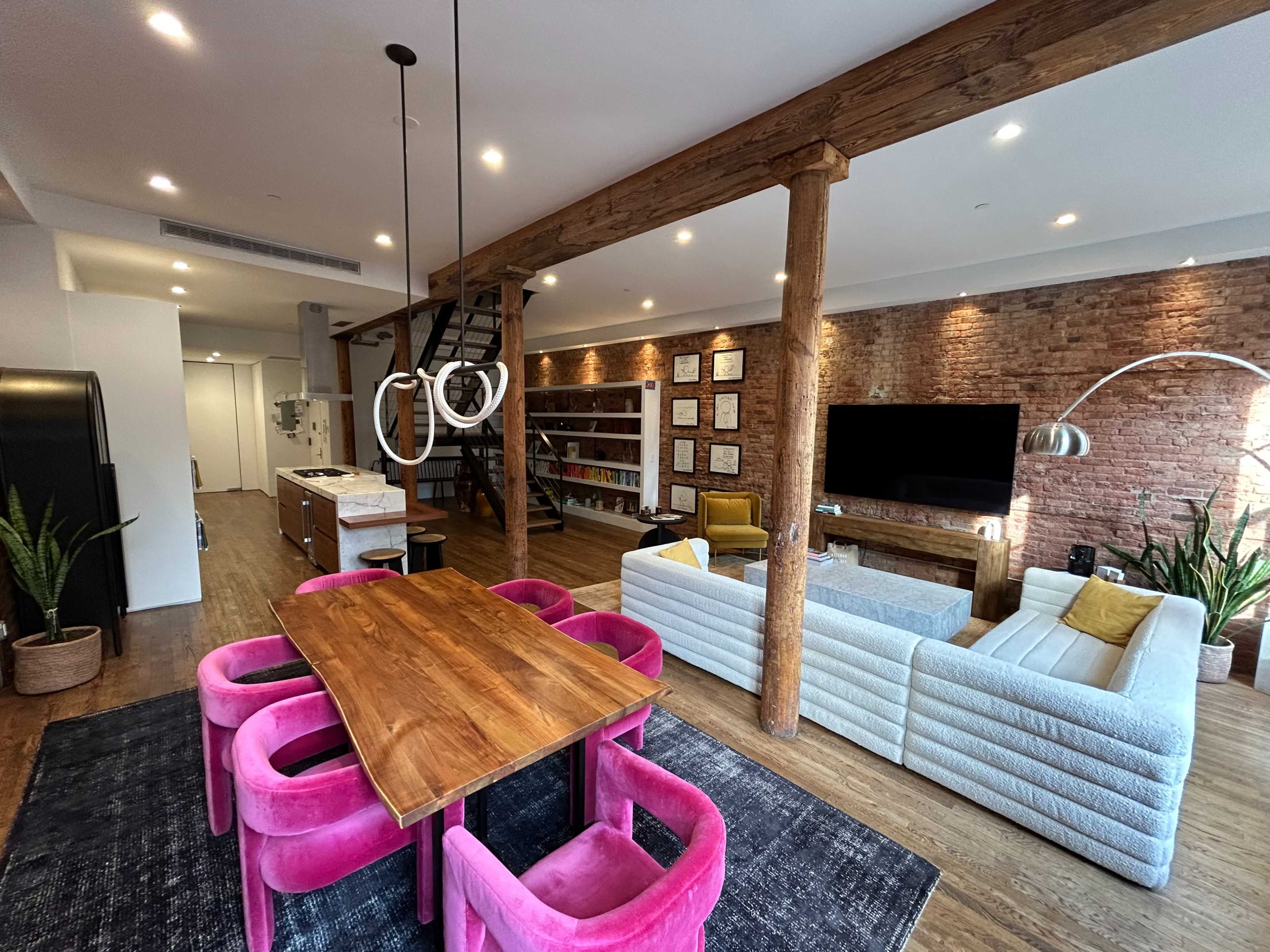 The image shows a modern open-concept living space featuring a dining area with a pink table and chairs, a cozy living area with sofas, and wooden beams supporting the ceiling.