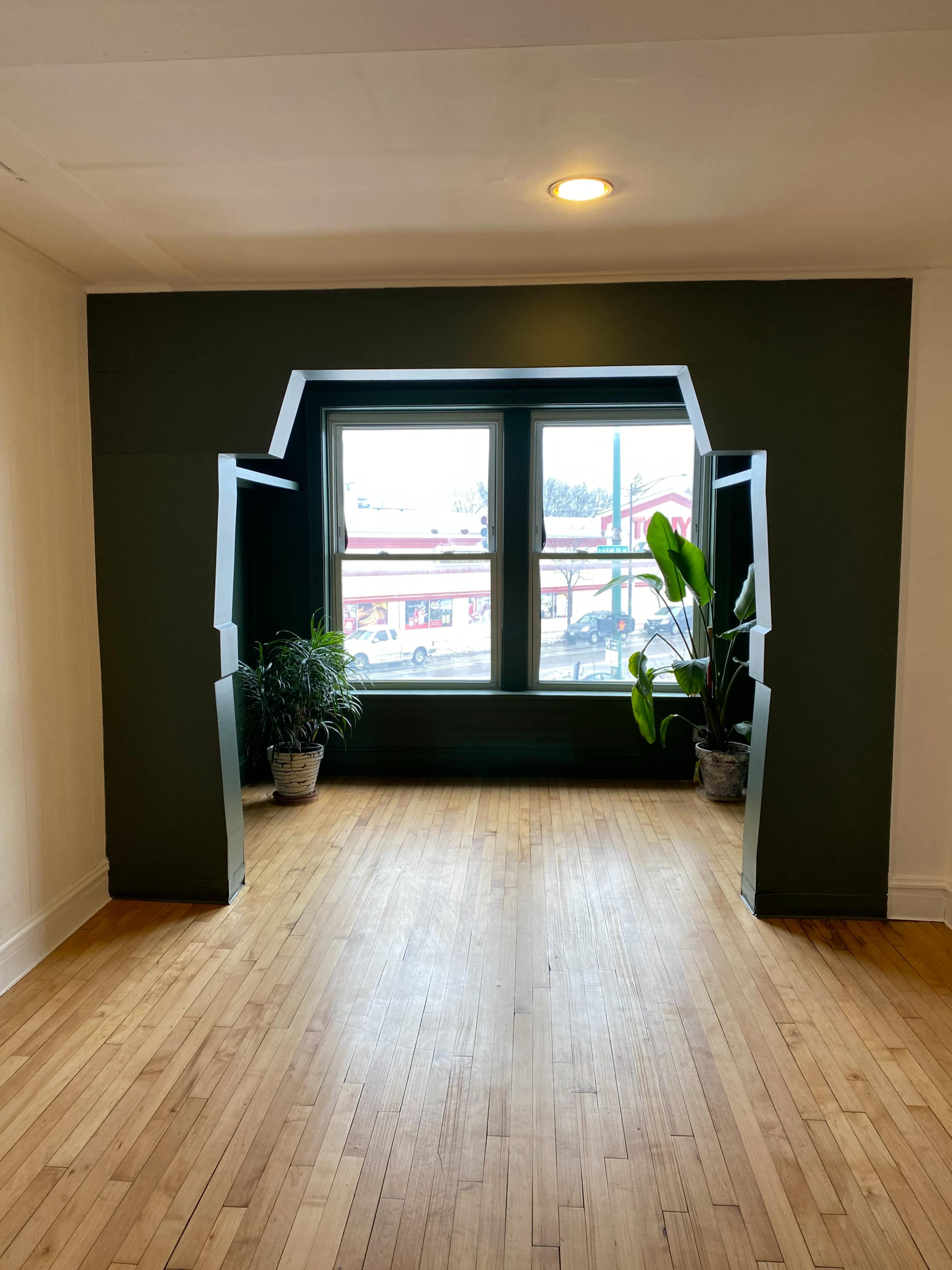 A room with wooden flooring and a large window framed by dark green architectural elements, flanked by potted plants.