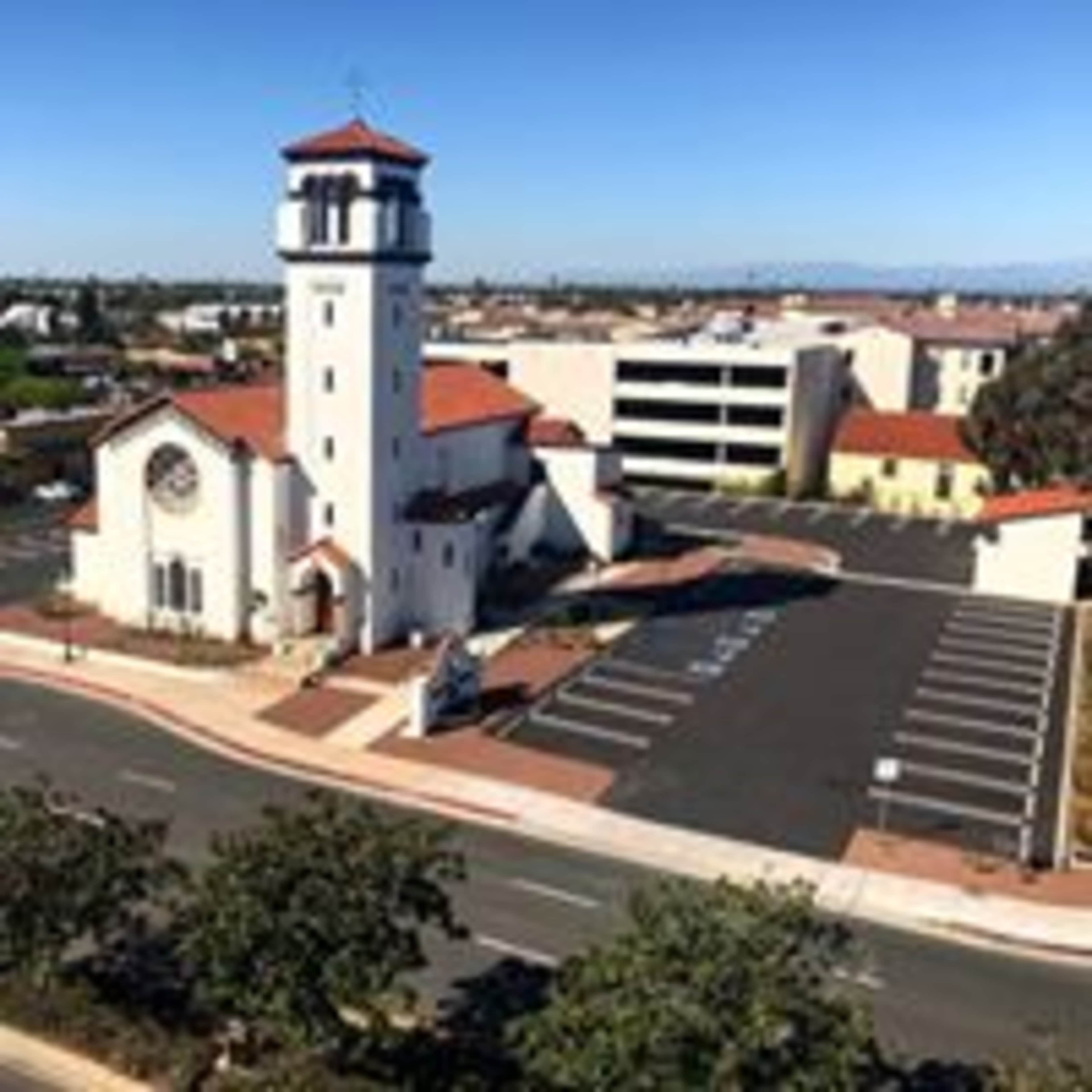 A church with a tall tower and red-tiled roof, situated next to a parking area and surrounded by trees and buildings.