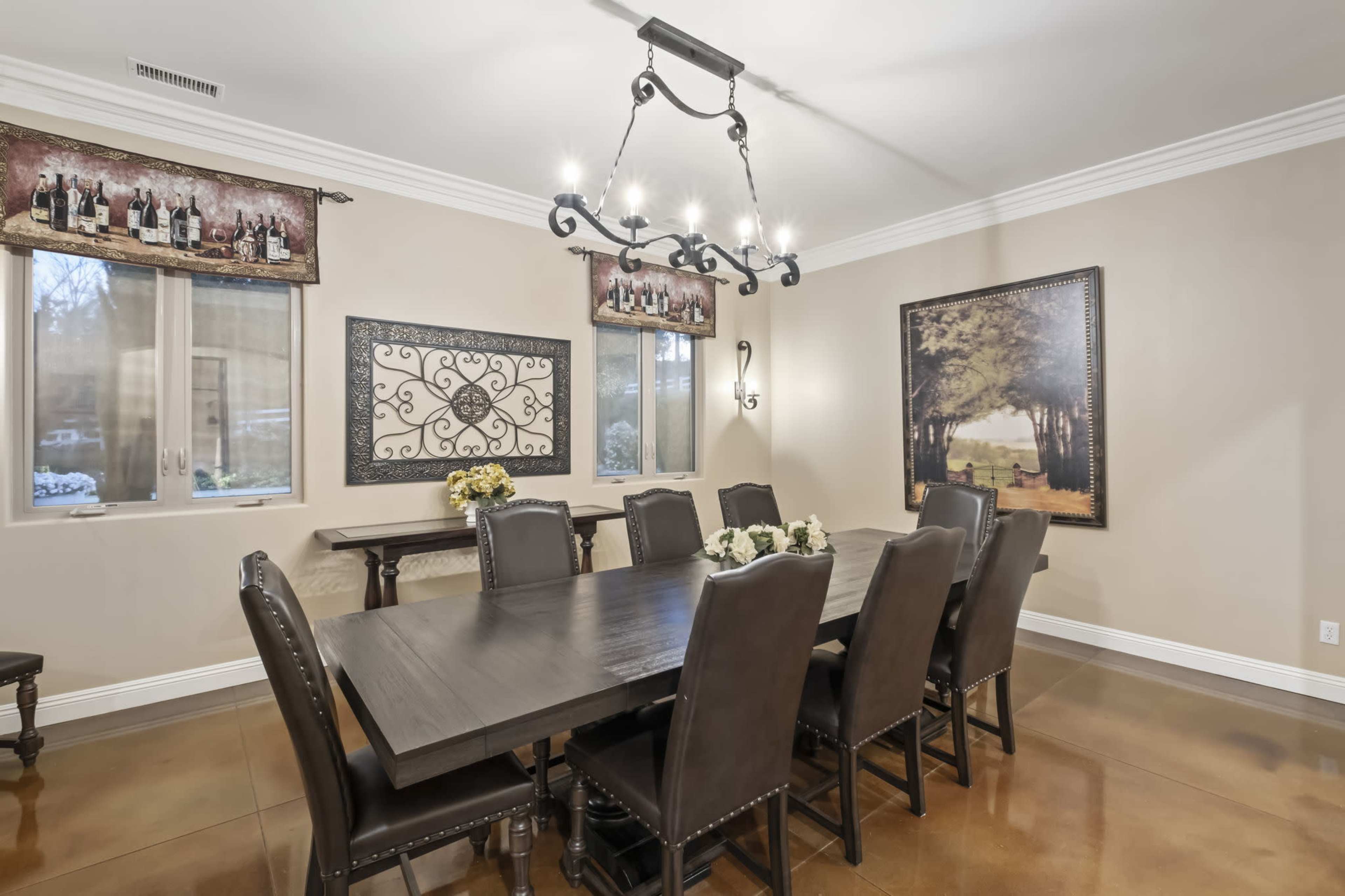 A dining room features a large wooden table surrounded by eight black chairs, with two windows and decorative artwork on the walls.