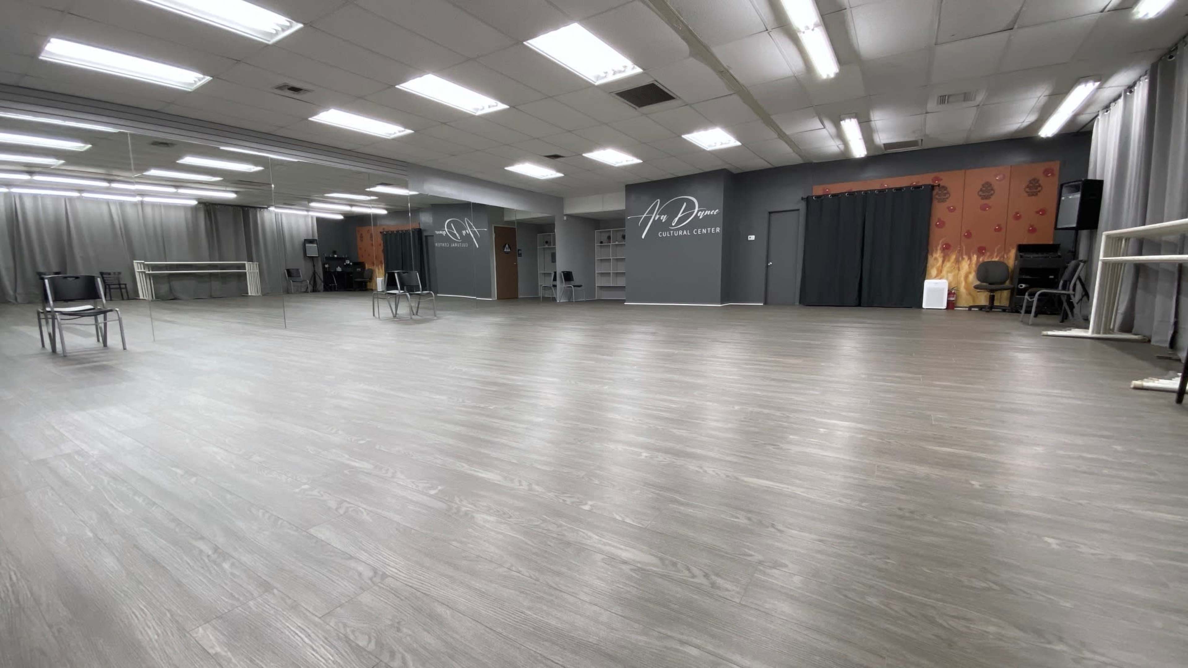 A spacious dance studio with wooden flooring, large mirrors, and a simple, uncluttered arrangement of chairs.