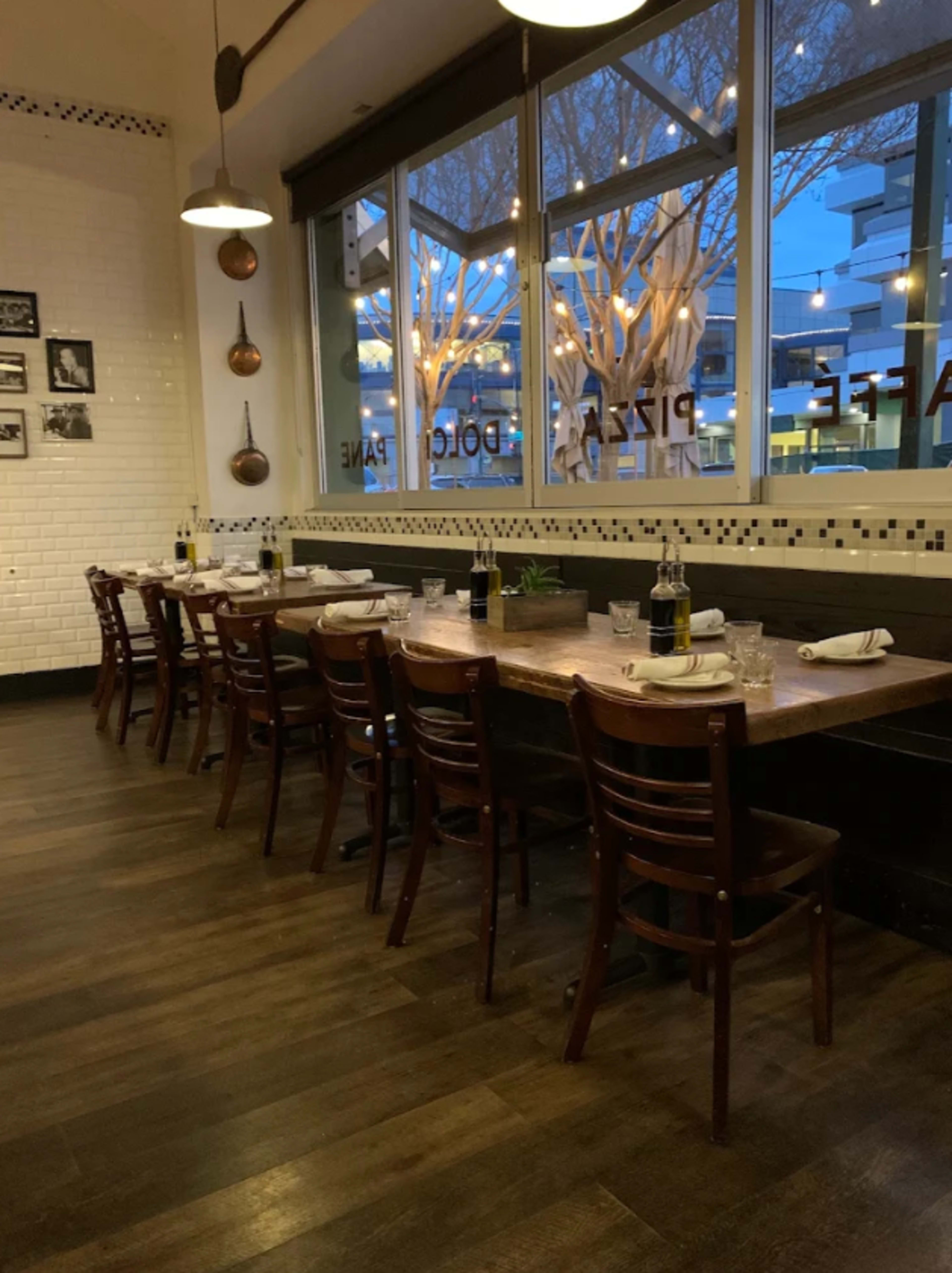 A long wooden table with chairs is set for dining in a restaurant, with large windows showing a twilight scene outside.