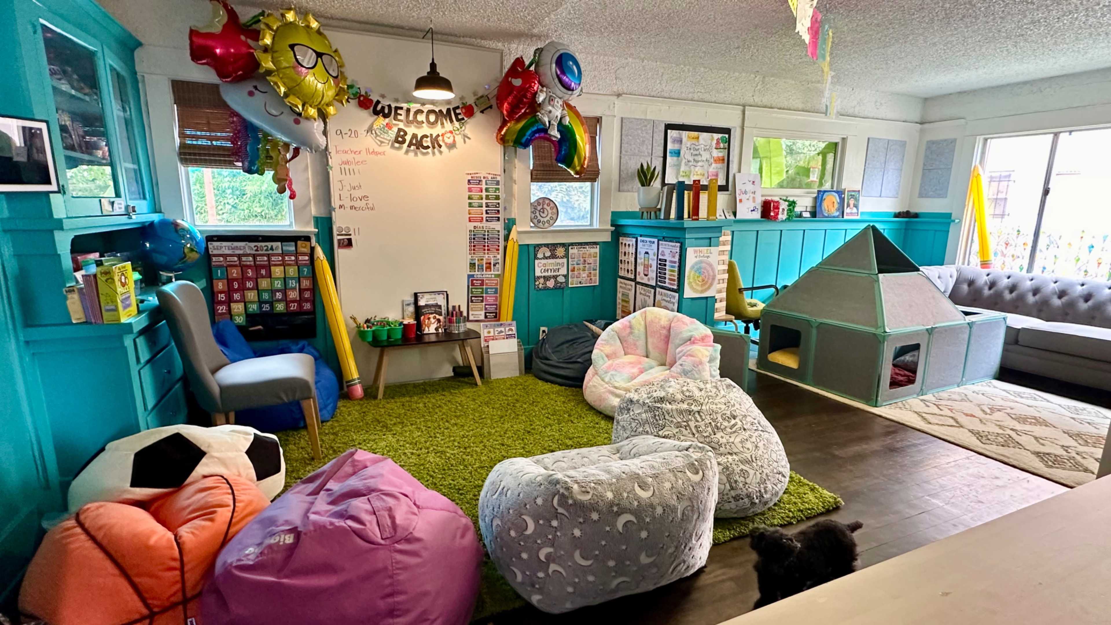 A colorful classroom setting features bean bag chairs, a play tent, and decorations including balloons and a welcome sign, along with a whiteboard and educational posters on the walls.