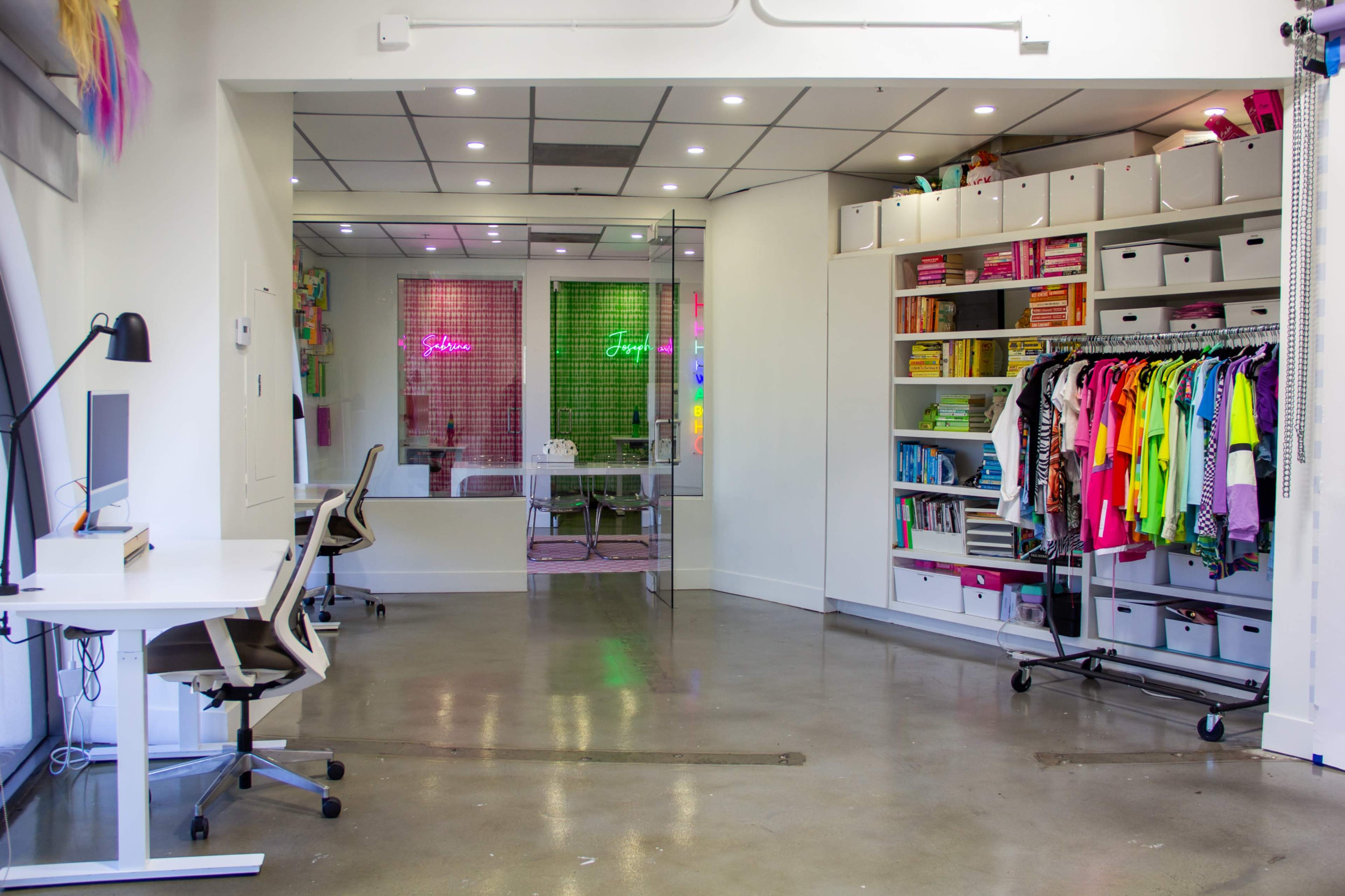 The image shows a bright, modern workspace featuring white desks, a colorful clothing rack, shelves filled with neatly arranged items, and vibrant neon signs in a glass-walled meeting area.