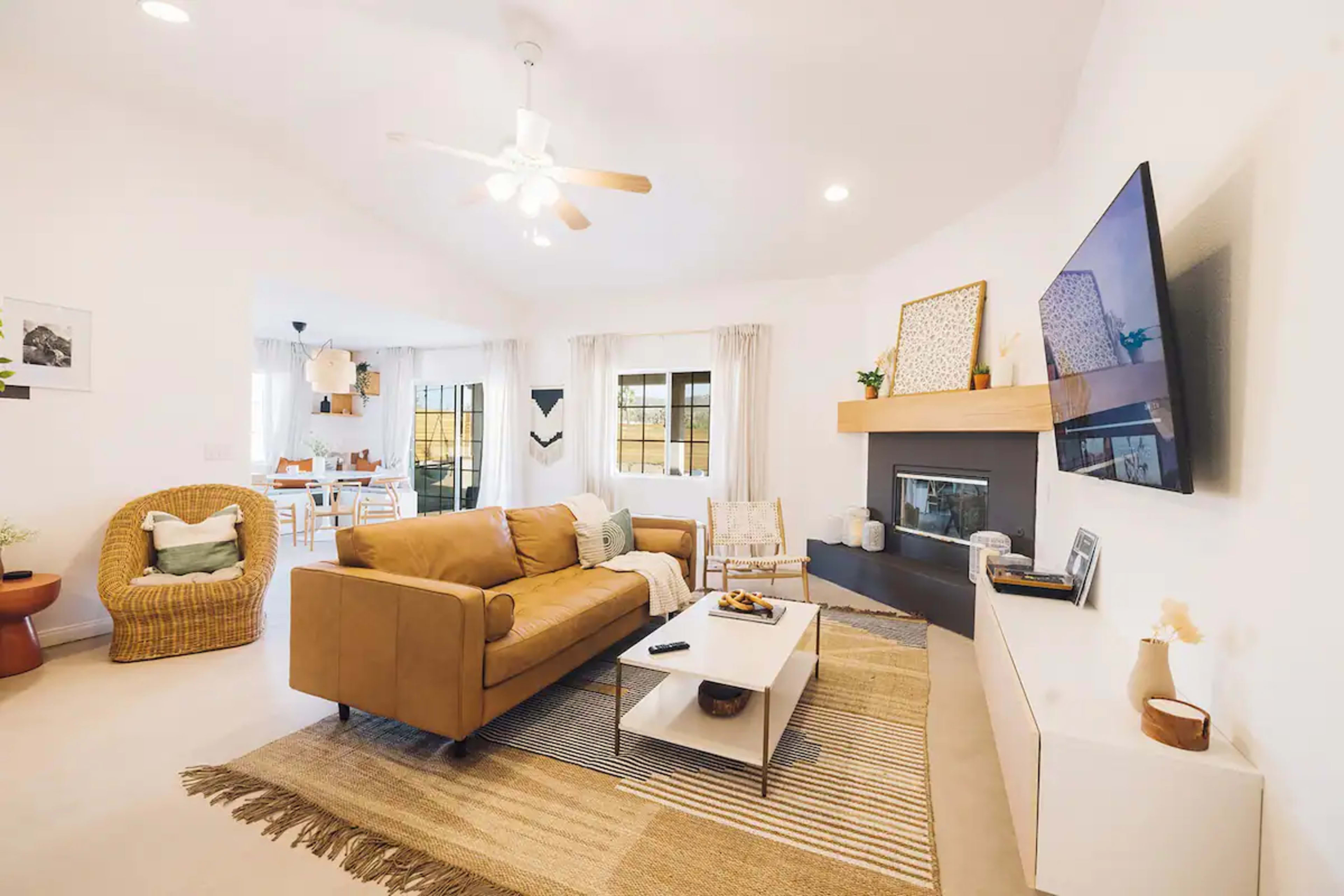 The living room features a tan sofa, a coffee table with snacks, and a wall-mounted television above a modern fireplace, with a dining area visible in the background.