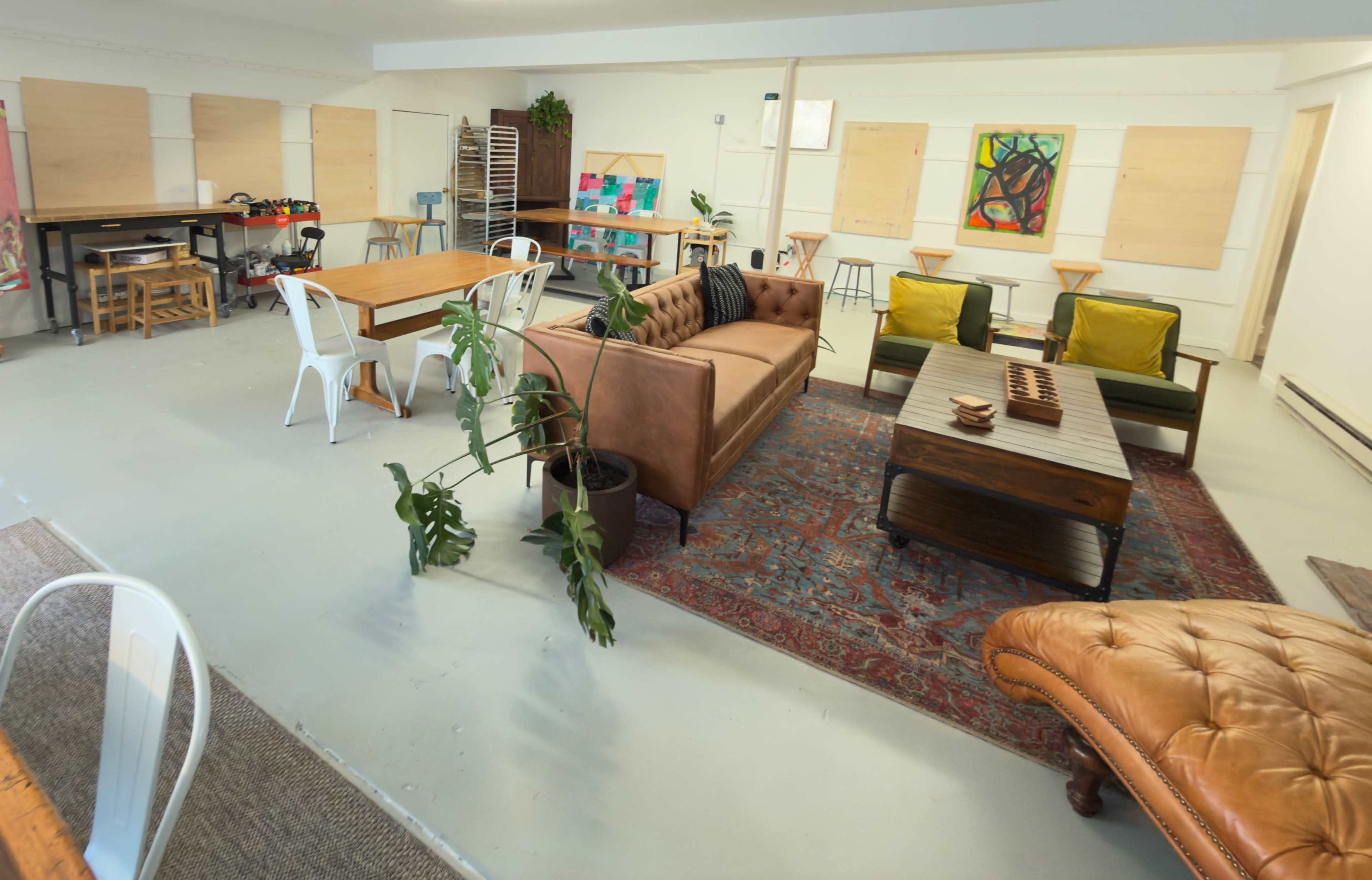 The room features a casual seating area with a brown leather sofa, two green chairs, a wooden coffee table, and a large rug, along with a dining table and various workstations.