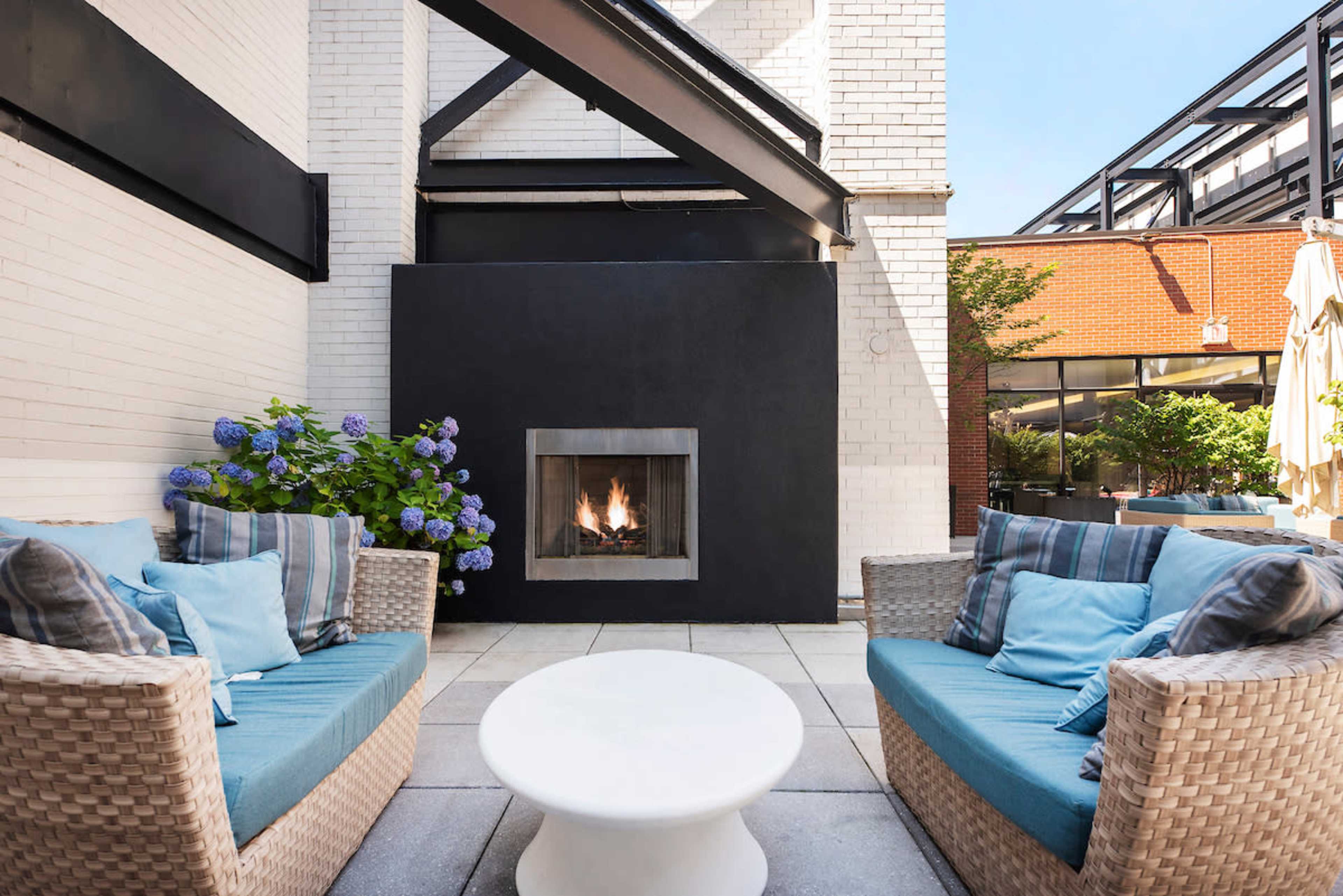 The image shows a modern outdoor patio with two wicker chairs, a round white coffee table, and a black fireplace surrounded by hydrangea plants.