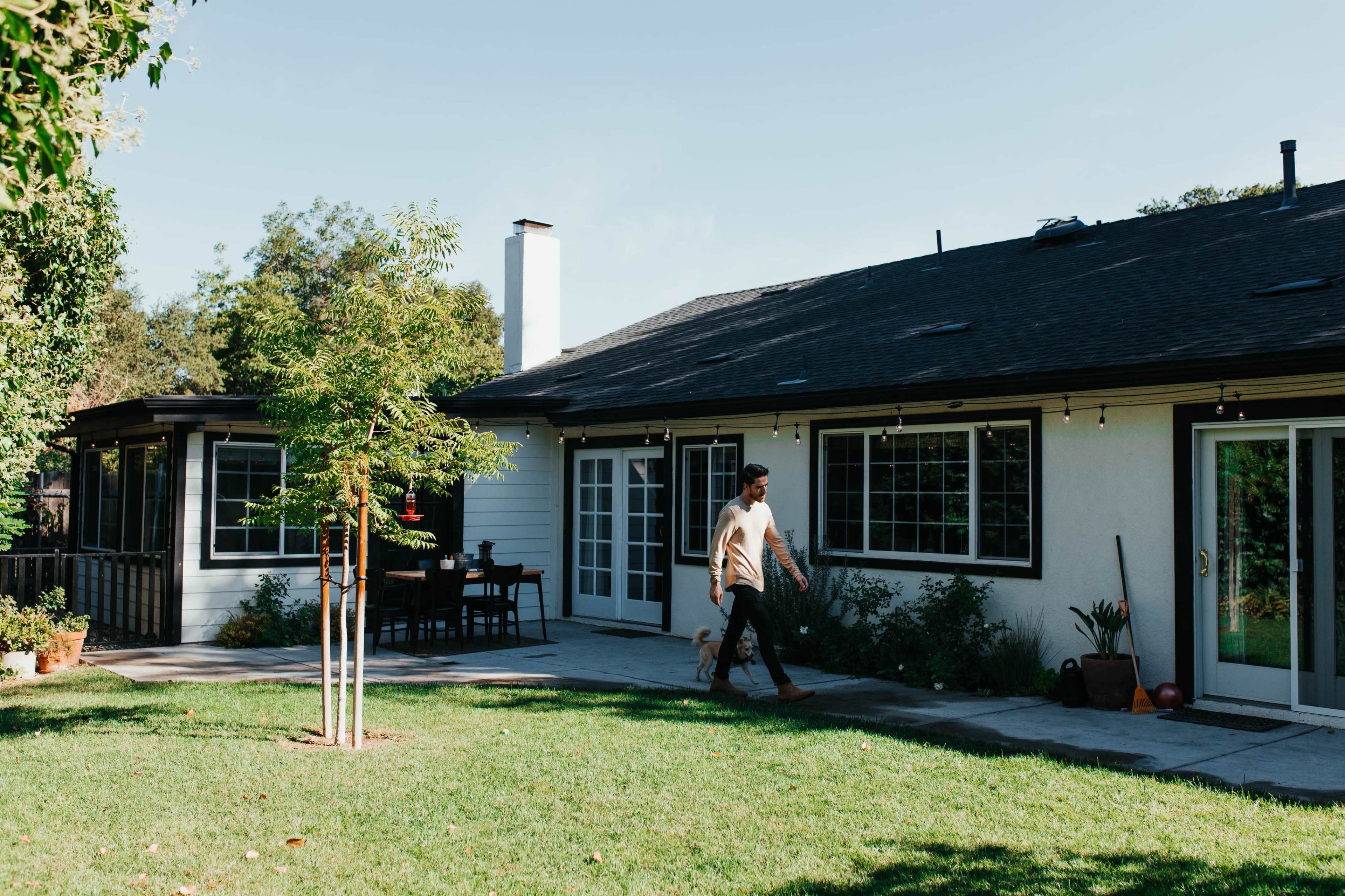 A man walks a dog across a grassy area in front of a single-story house with a patio and large windows.