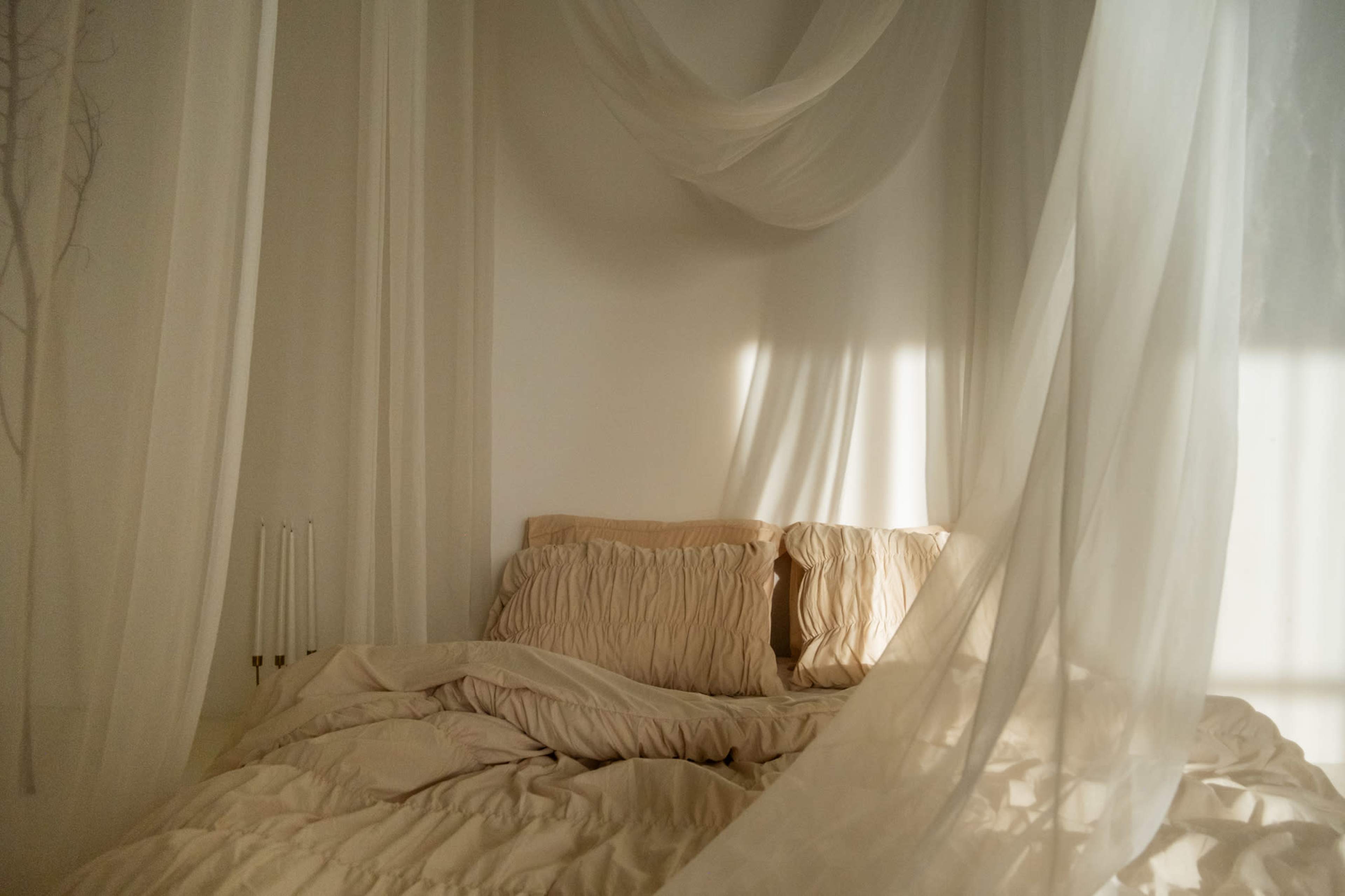 A neatly arranged bed with soft beige bedding and pillows is surrounded by flowing sheer curtains that softly filter the light.