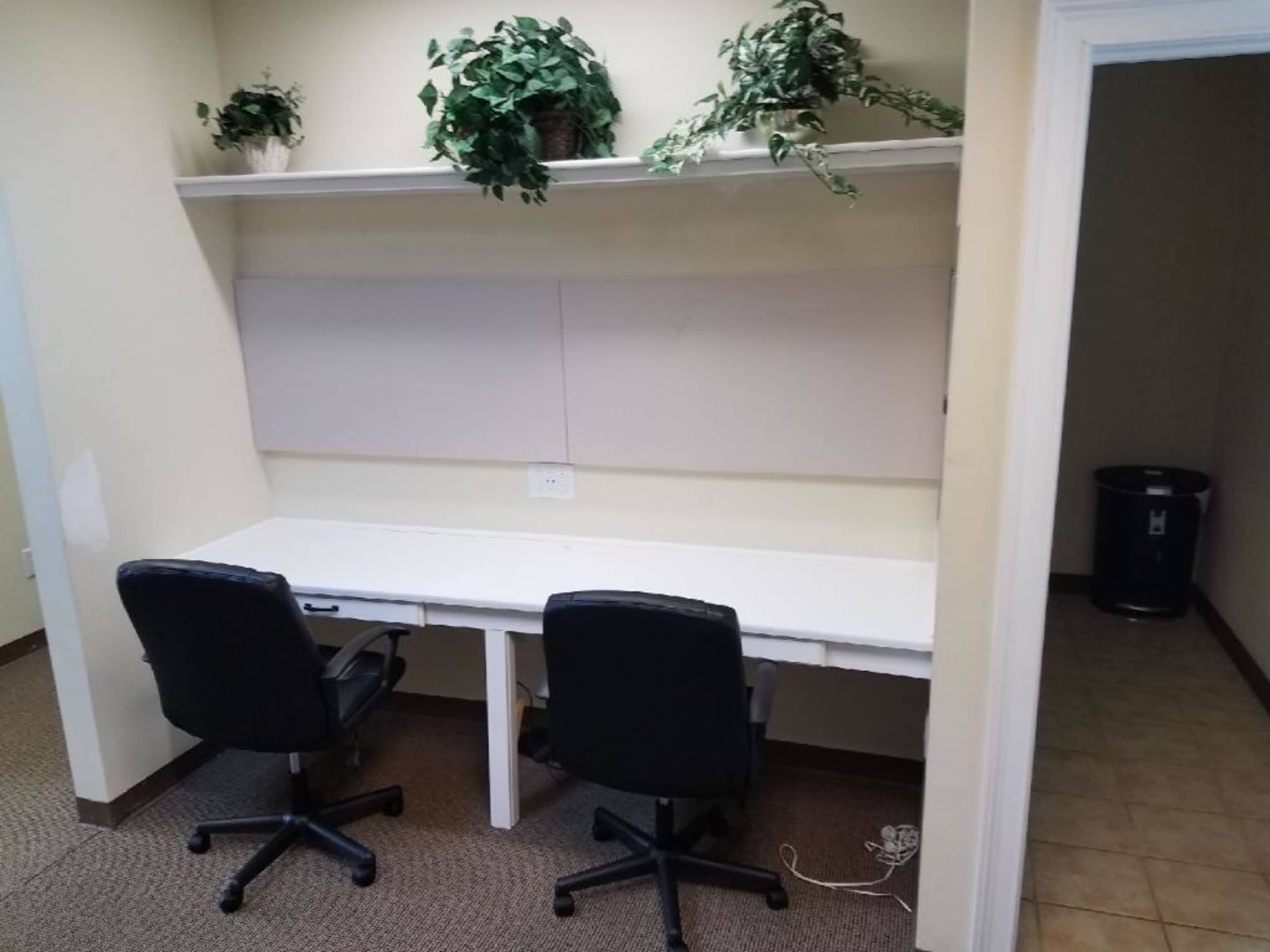 A small office space features two black chairs positioned at a white desk with plants on a shelf above and a door leading to another room on the right.