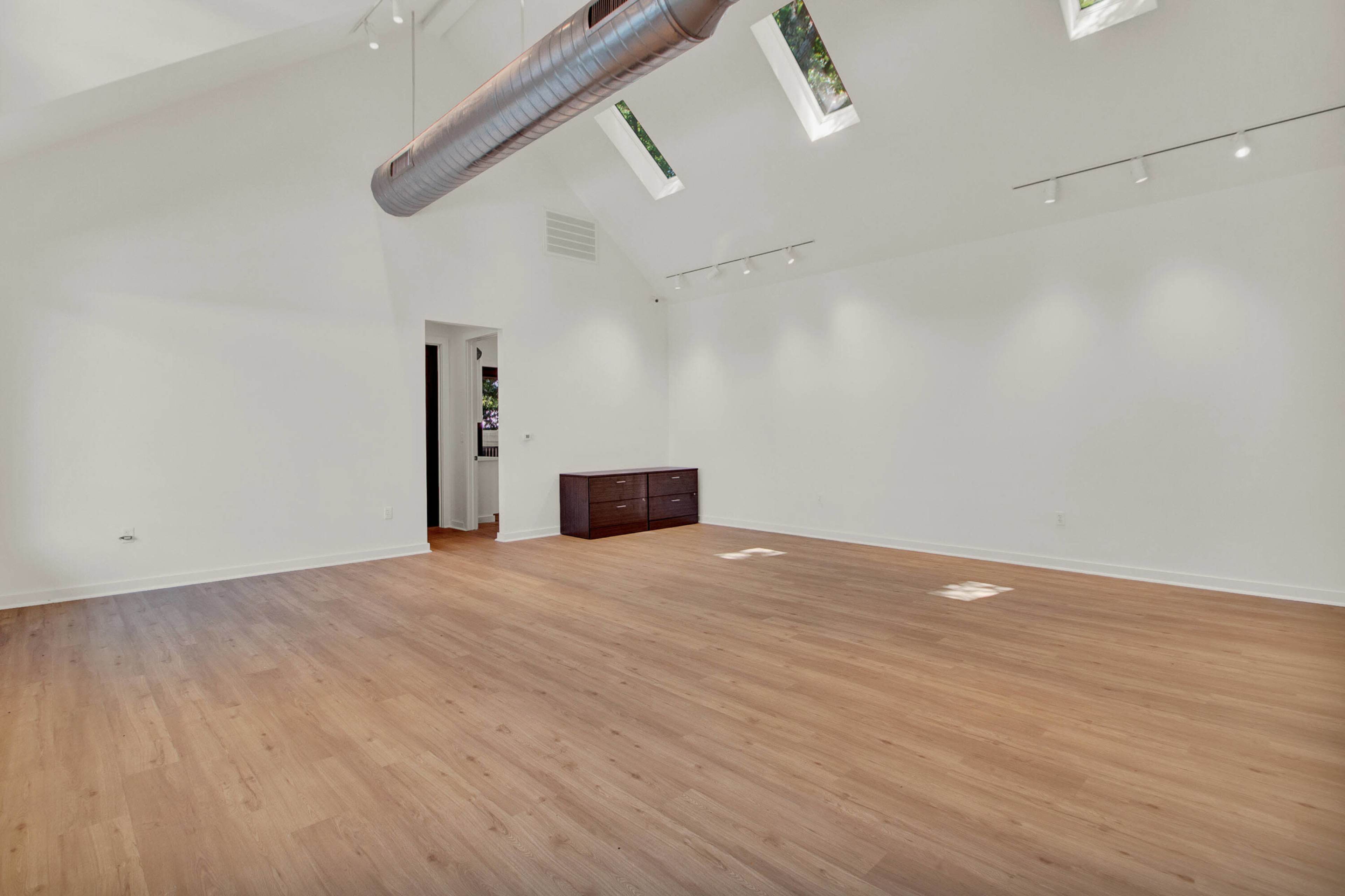 The image shows a spacious, empty room with wooden flooring, high ceilings, and several skylights allowing natural light to enter.