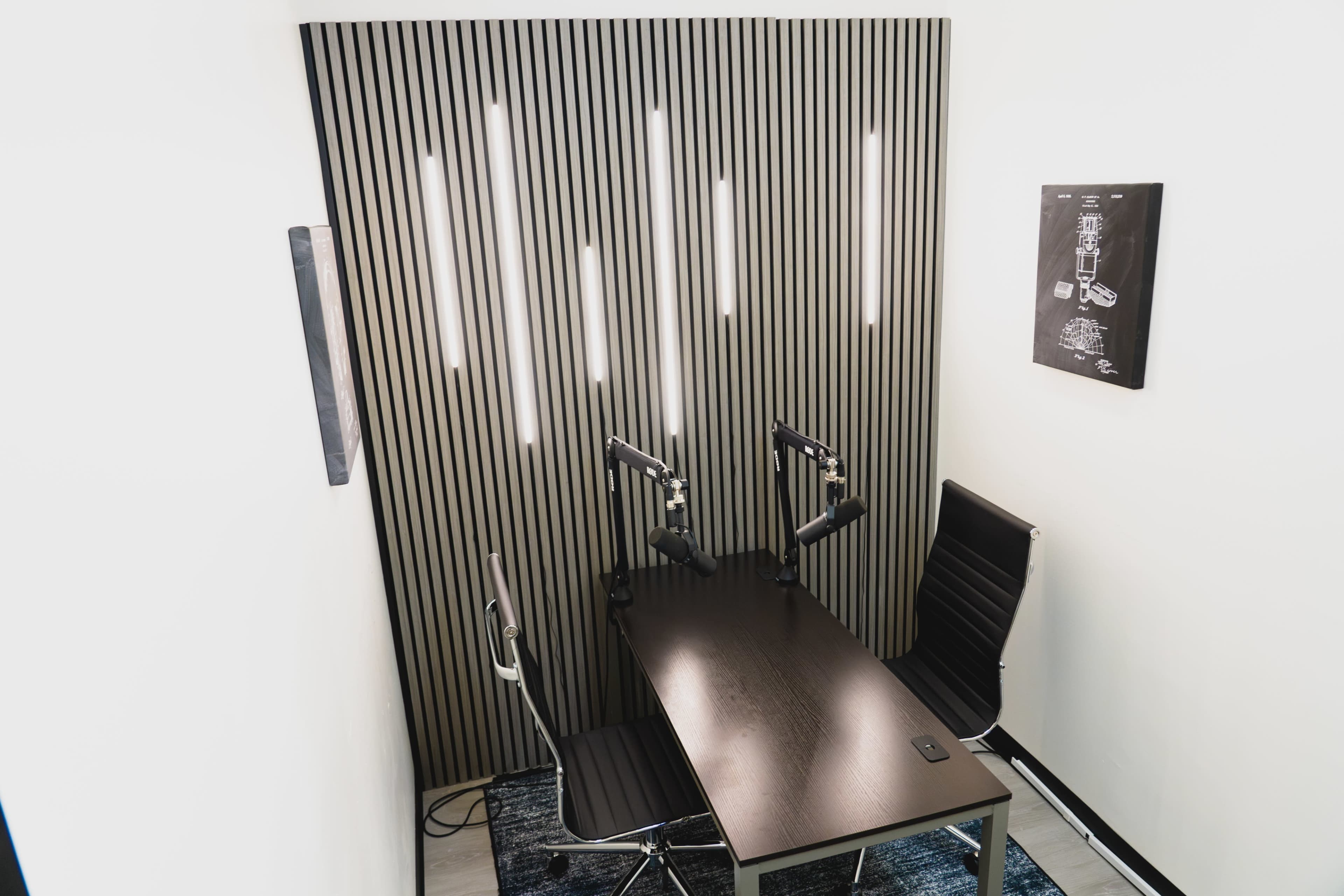 The image shows a small recording studio with a wooden table, two chairs, and a slatted wall featuring overhead lights.