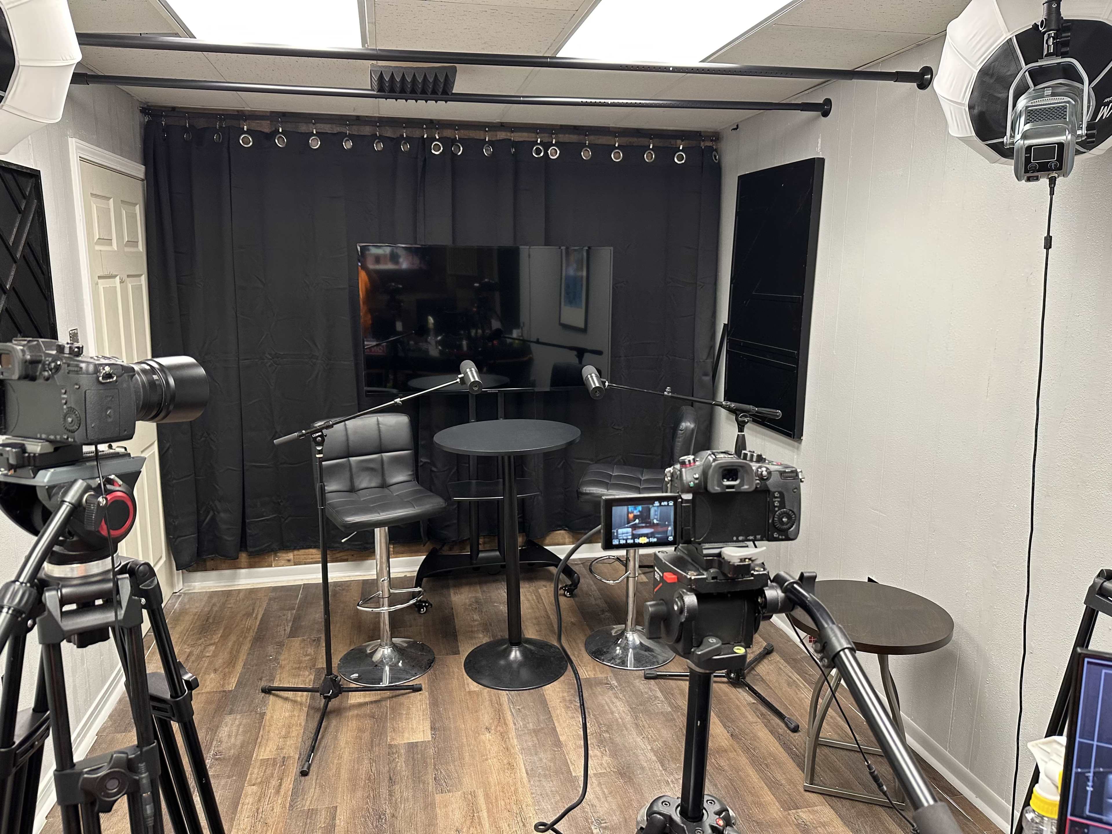 The image shows a small video recording setup featuring two chairs, two microphones on stands, a large television, and several cameras positioned on tripods.