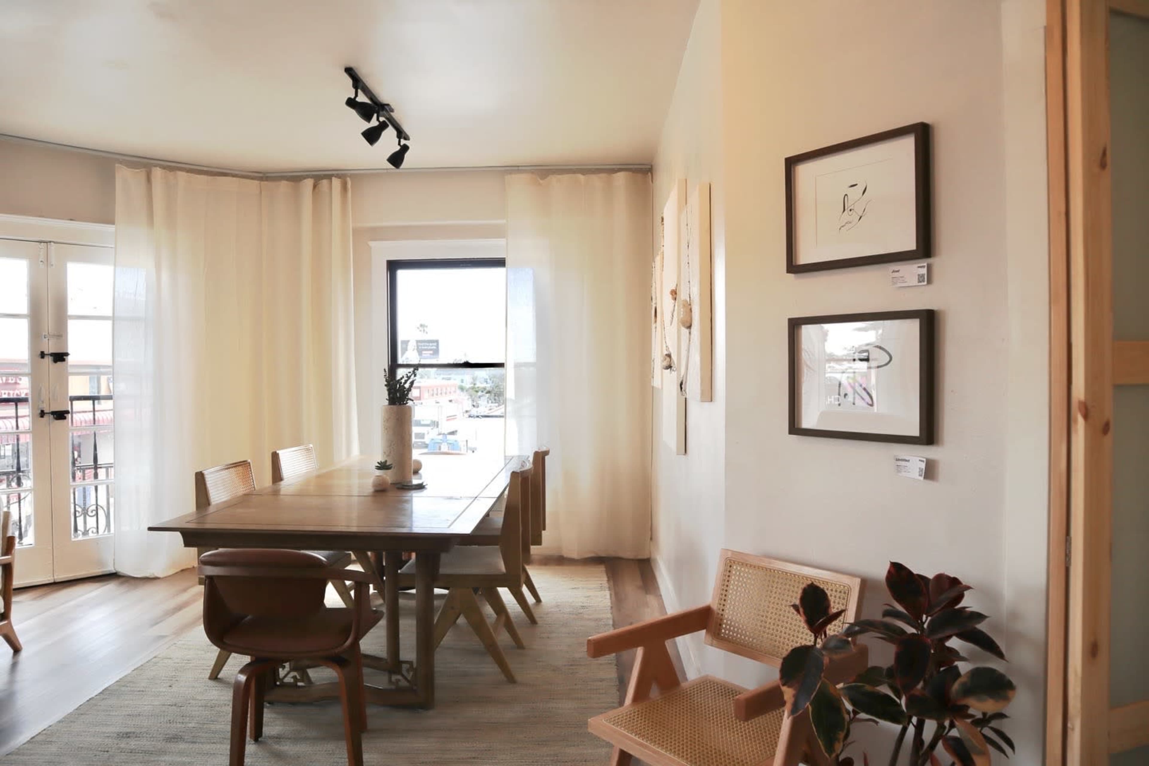 A dining area features a wooden table surrounded by chairs, with sheer curtains covering a window and wall art displayed on the walls.