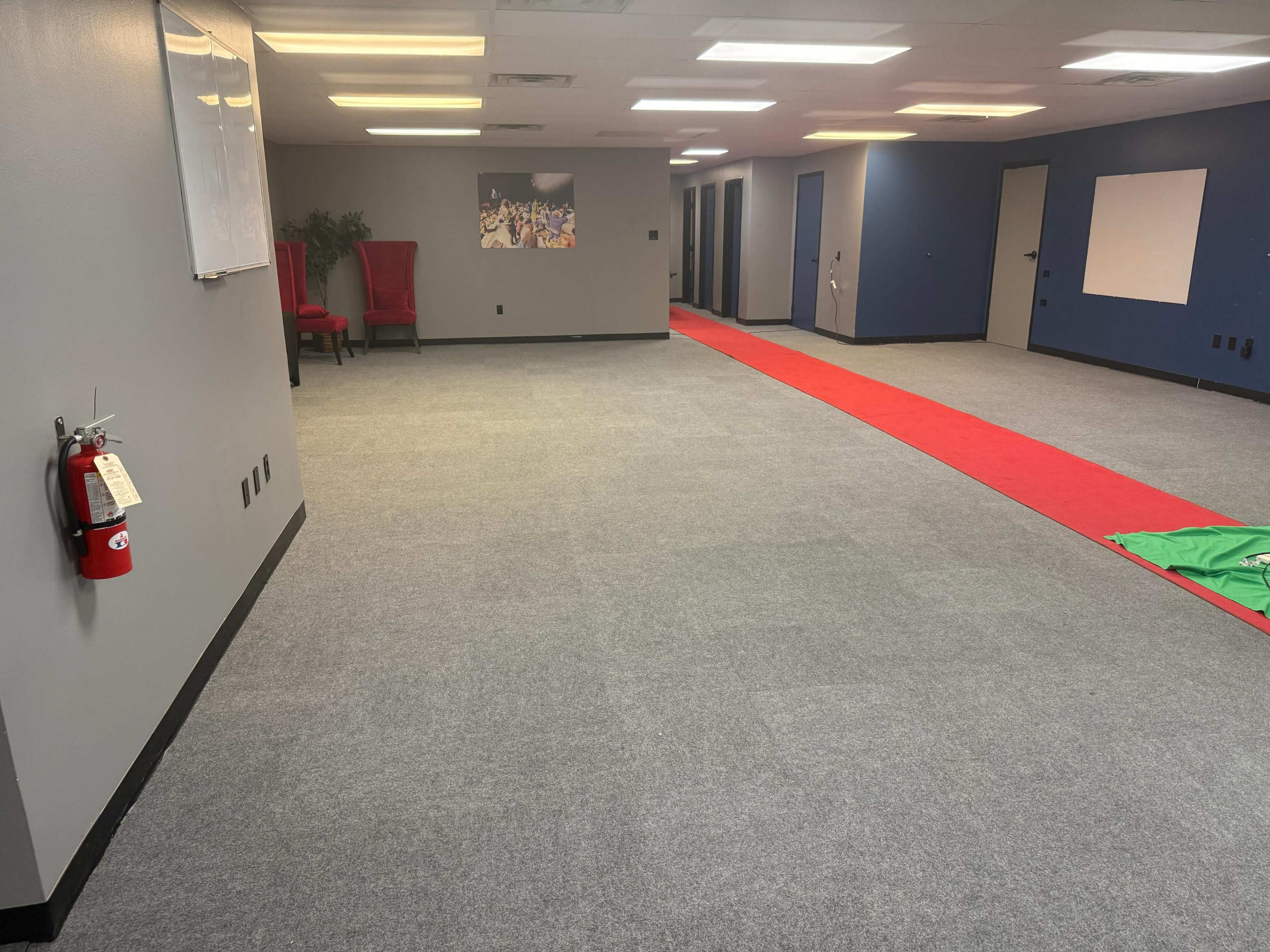 The image shows a spacious, empty room with gray carpet flooring, a red carpet laid out in the center, two red chairs against one wall, and light-colored walls with a few doors and a whiteboard.