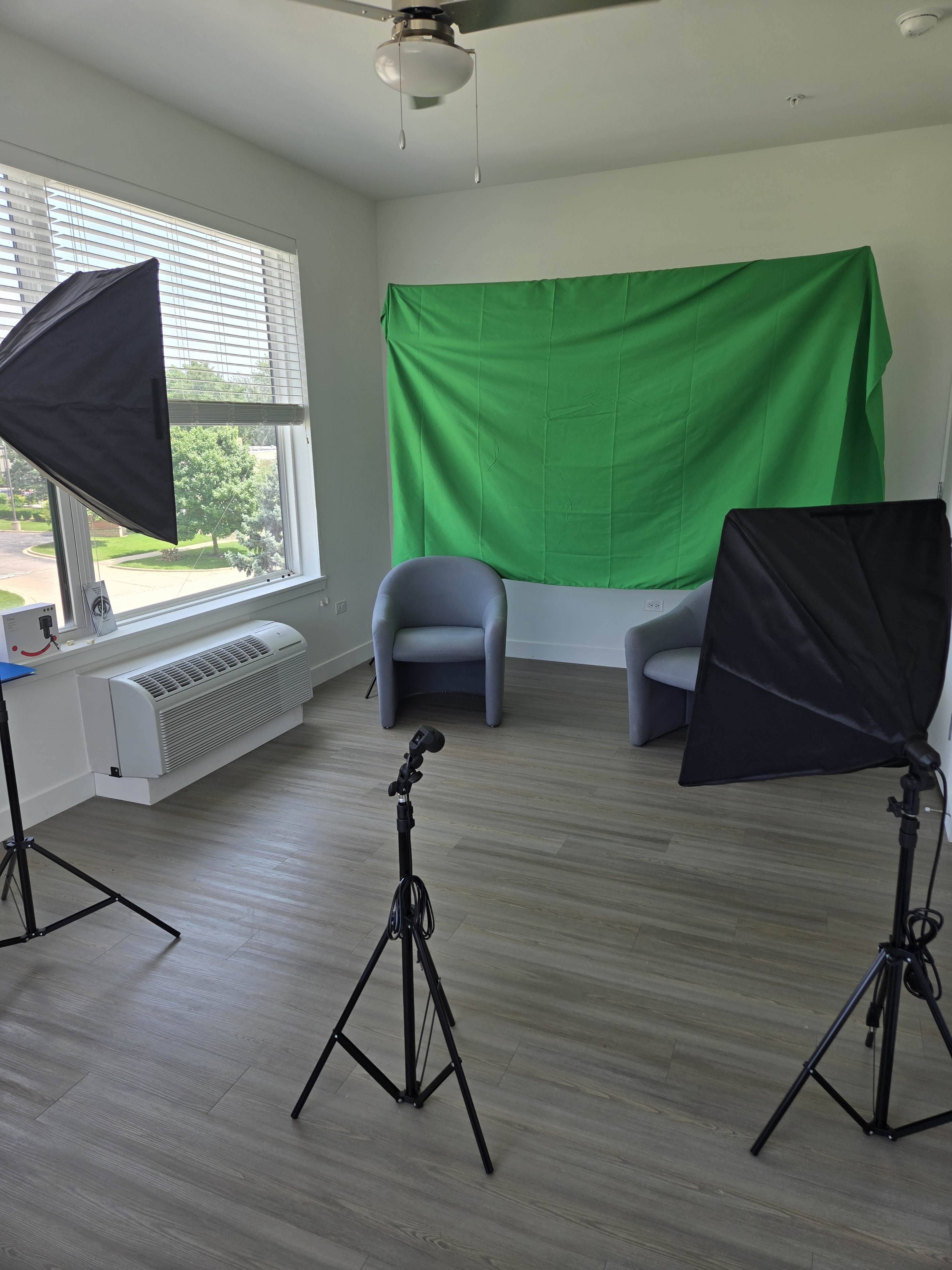The image shows a well-lit room with two chairs facing a green backdrop, flanked by two light stands on either side.