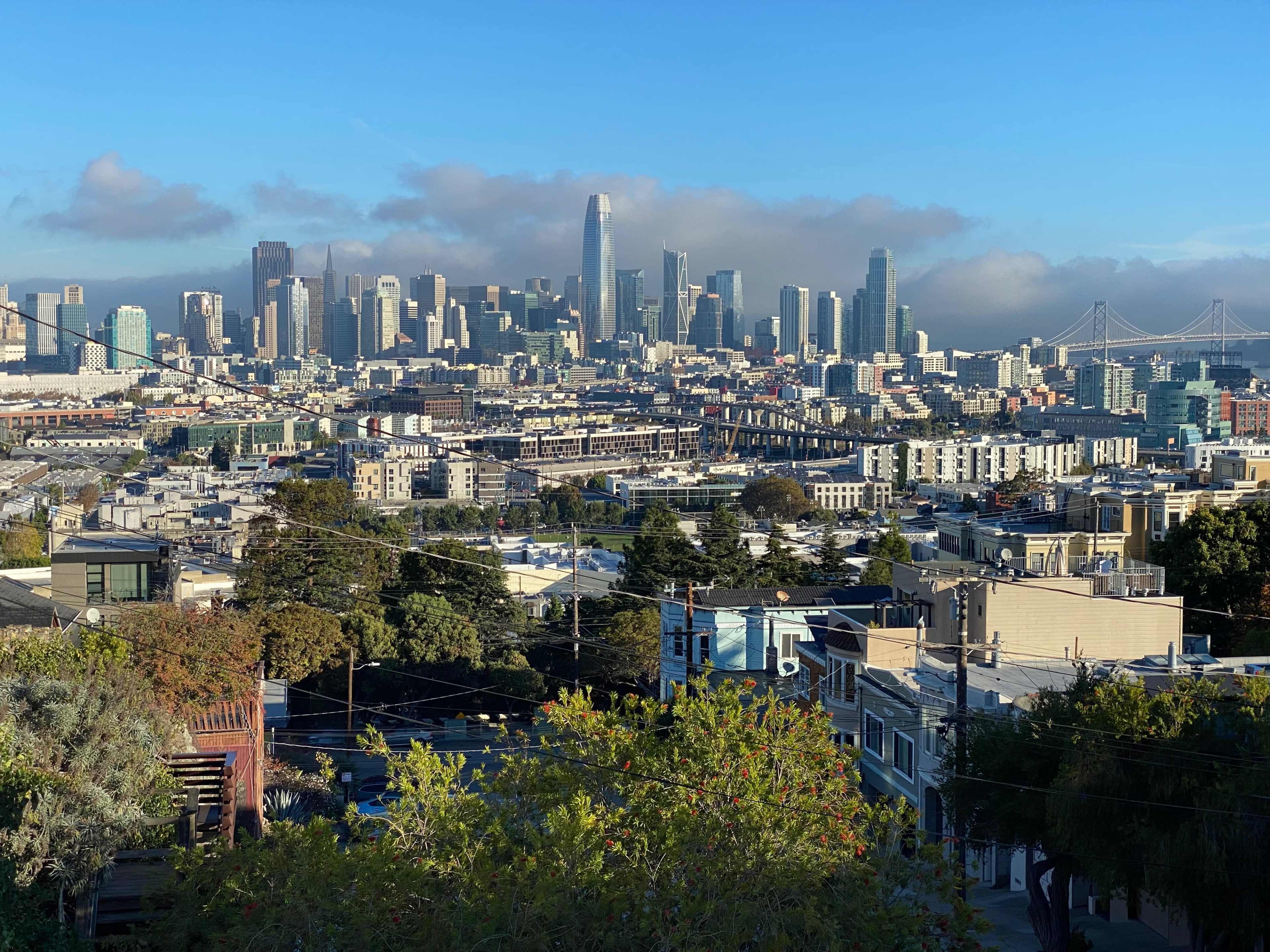 A panoramic view of the San Francisco skyline, featuring tall buildings and the Golden Gate Bridge in the background under a clear blue sky.