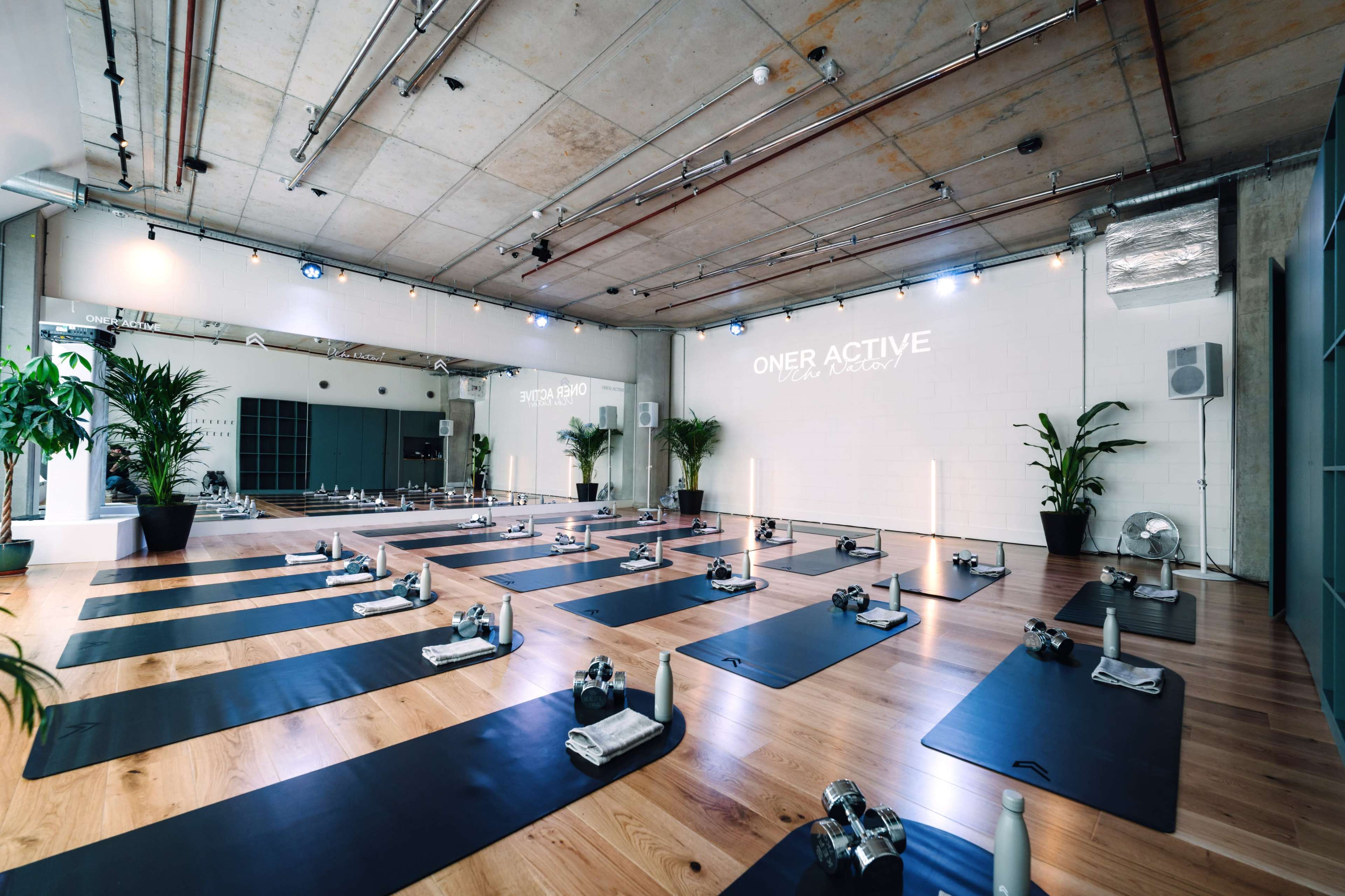 The image shows a spacious yoga studio with neatly arranged mats, water bottles, and potted plants, illuminated by soft overhead lighting.