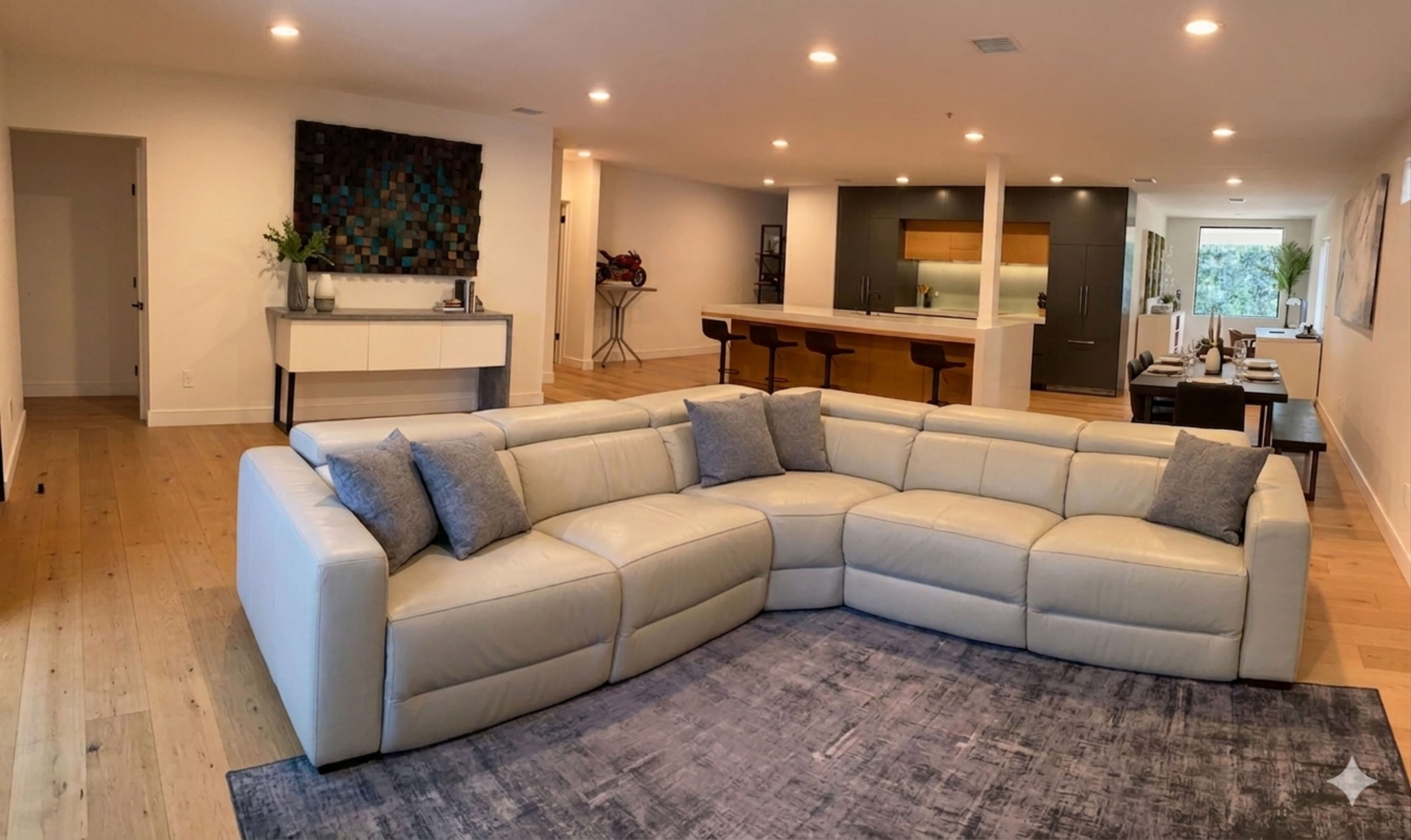 A modern living room features a spacious sectional leather sofa, a large area rug, and an open layout leading to a kitchen with bar seating.