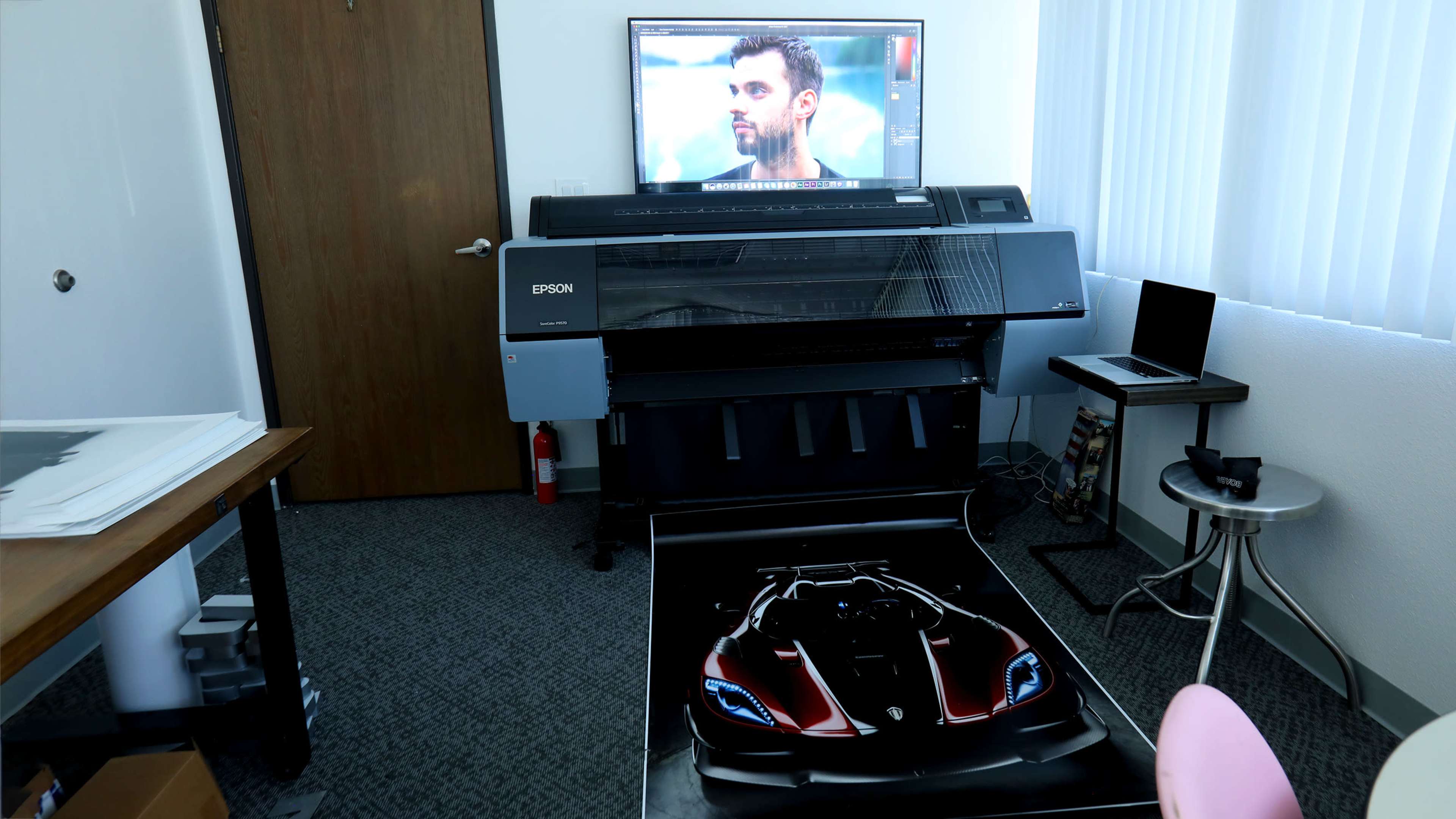 A large Epson printer is positioned in an office space, displaying a detailed image of a sports car on its output tray, with a television screen showing a man's face in the background.
