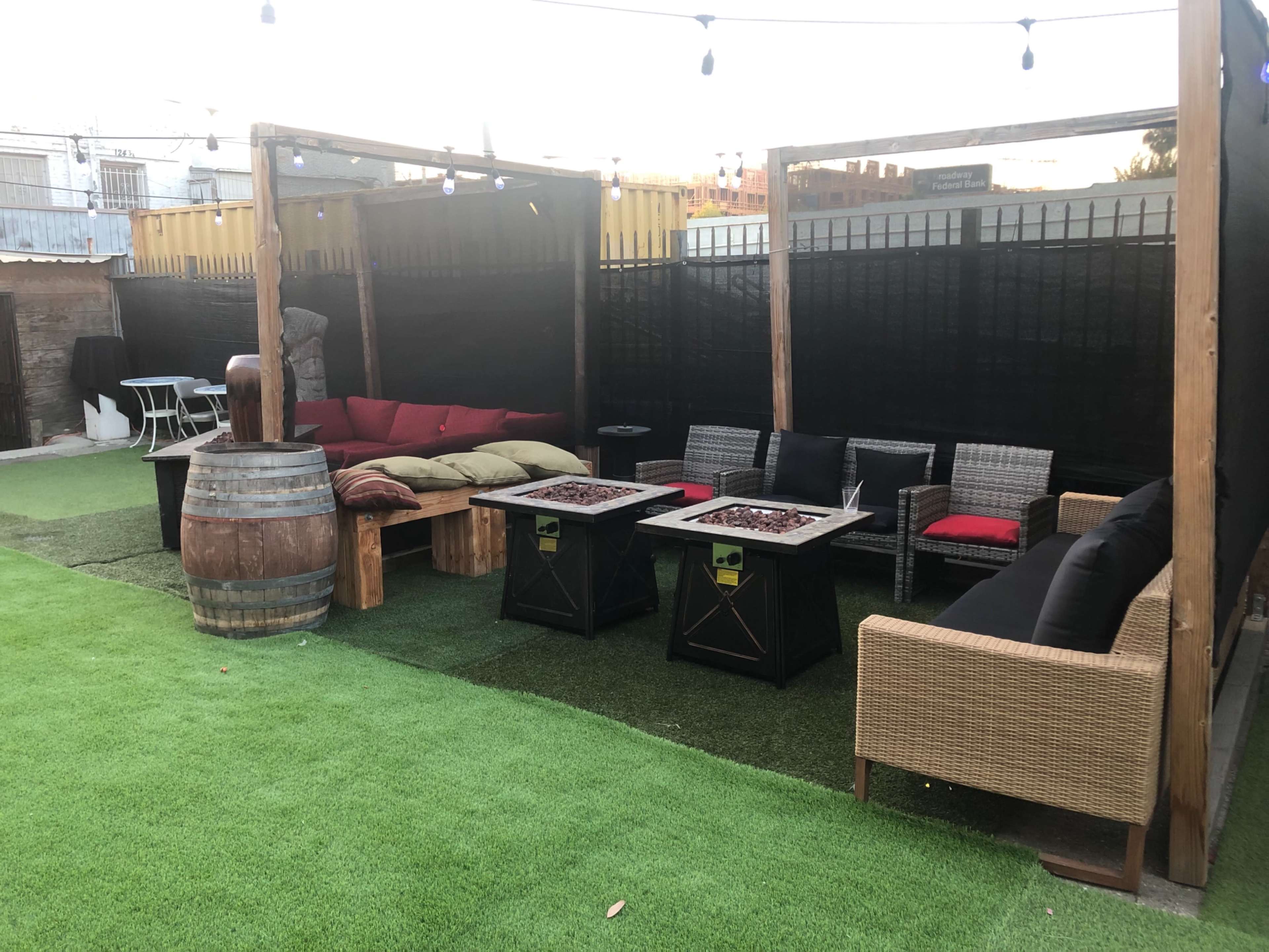 The image shows a backyard patio area with seating arrangements, including wooden benches, wicker sofas, and a barrel, surrounded by synthetic grass and string lights.