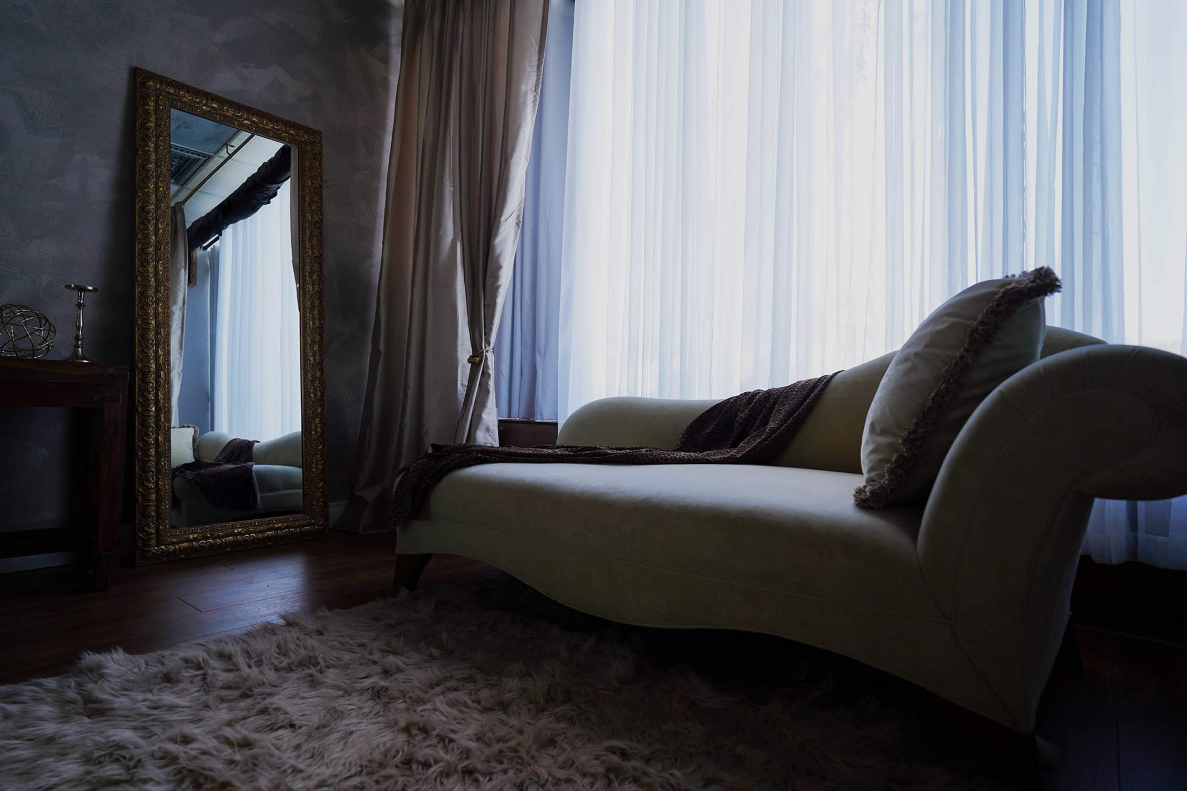 A light green chaise lounge is placed beside a large ornate mirror in a softly lit room with sheer curtains.