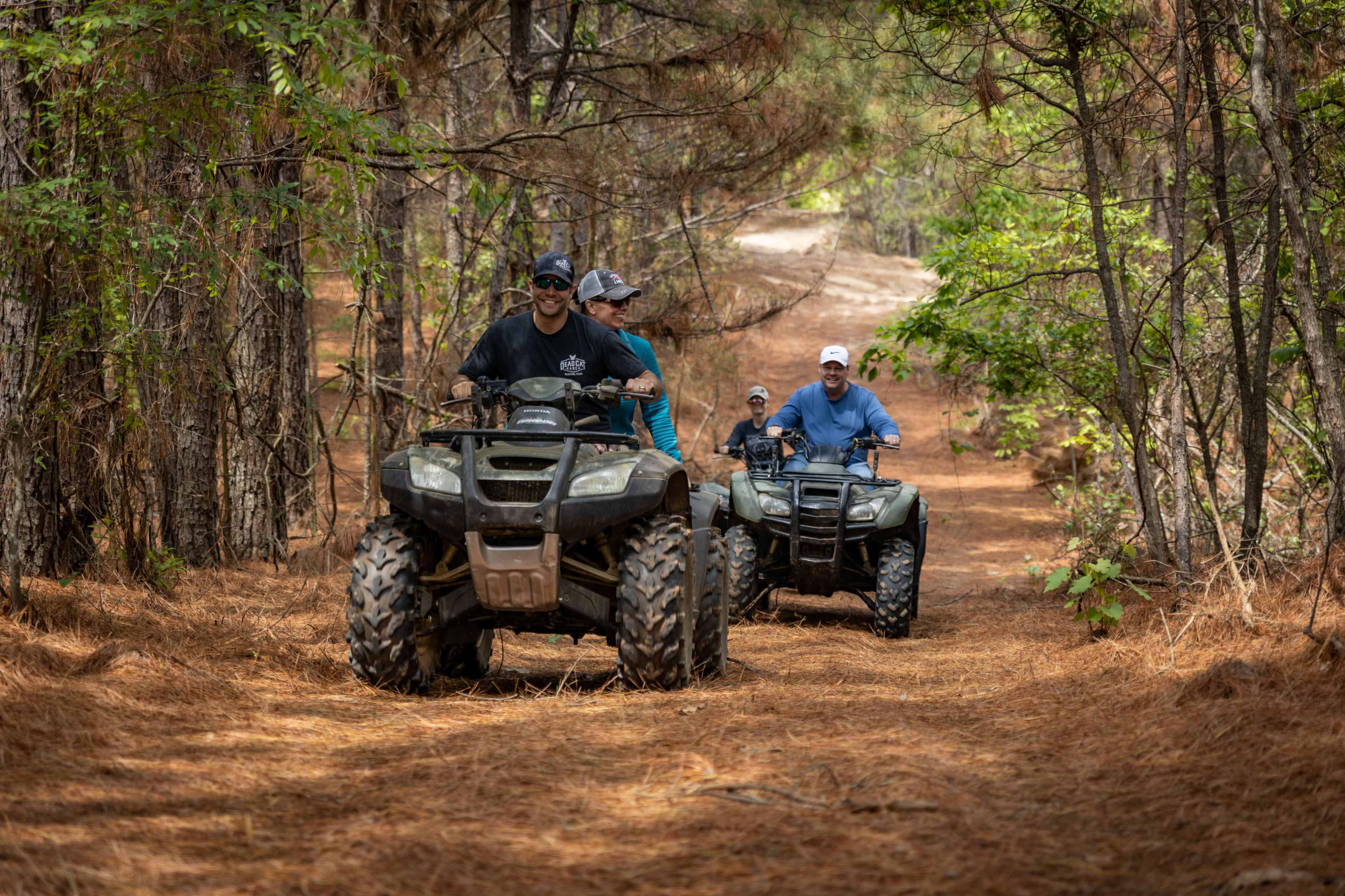 Two people ride all-terrain vehicles along a wooded path covered in pine needles.