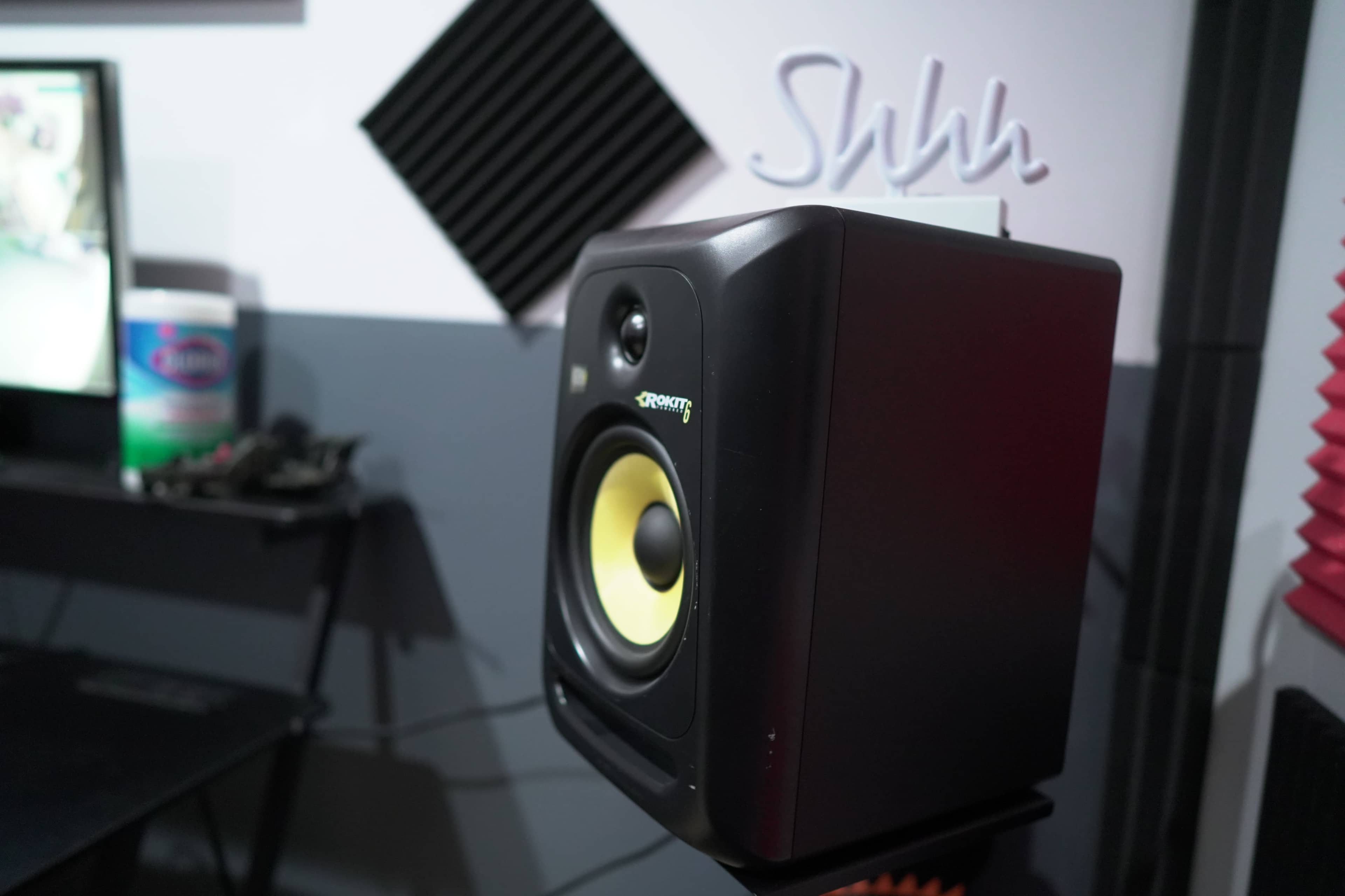 A black speaker with a yellow cone is positioned on a stand near a wall with soundproofing panels and a television in the background.
