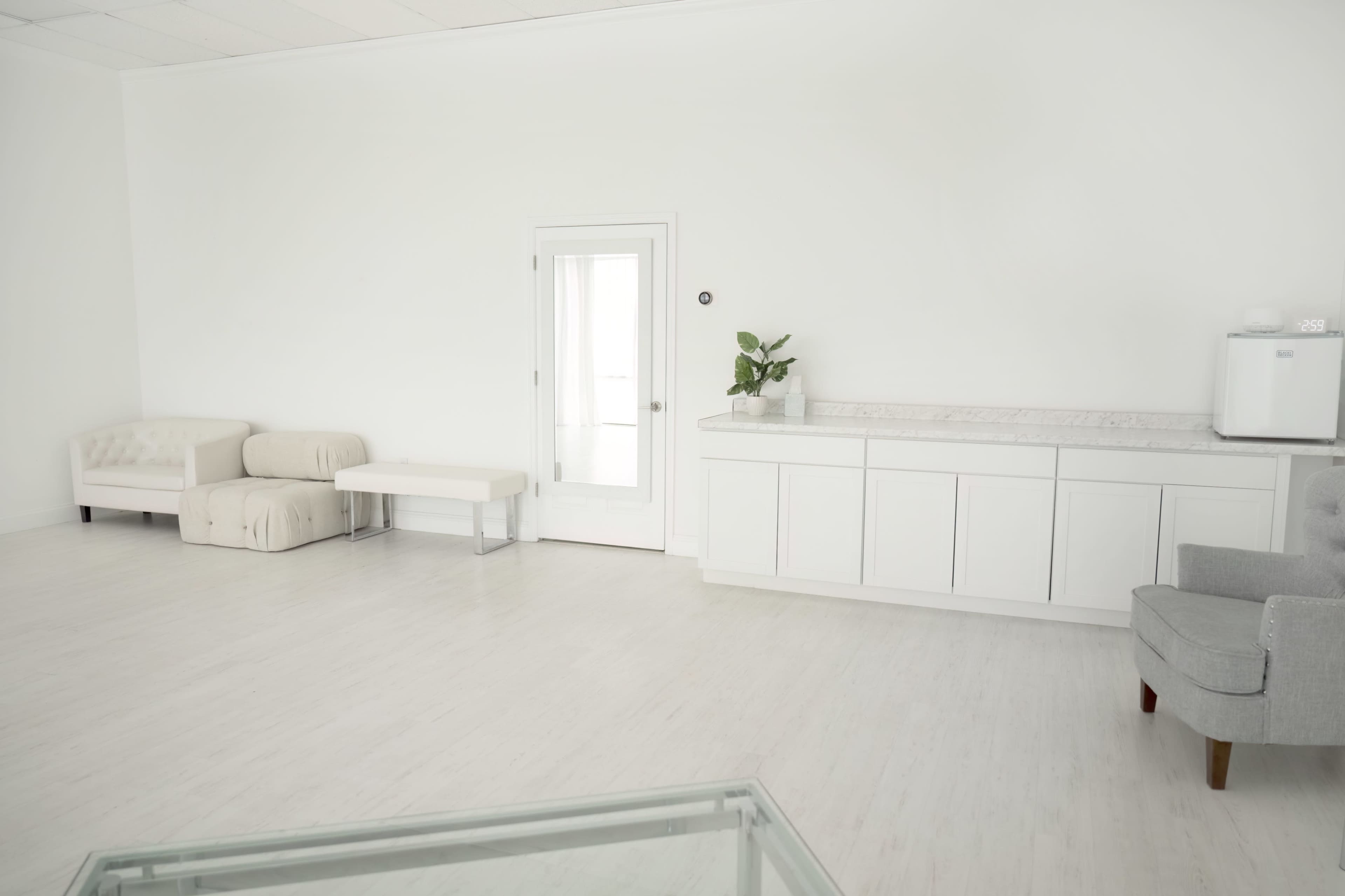The room features white walls and floors, with a sofa, a bench, a cabinet, and a door leading outside.