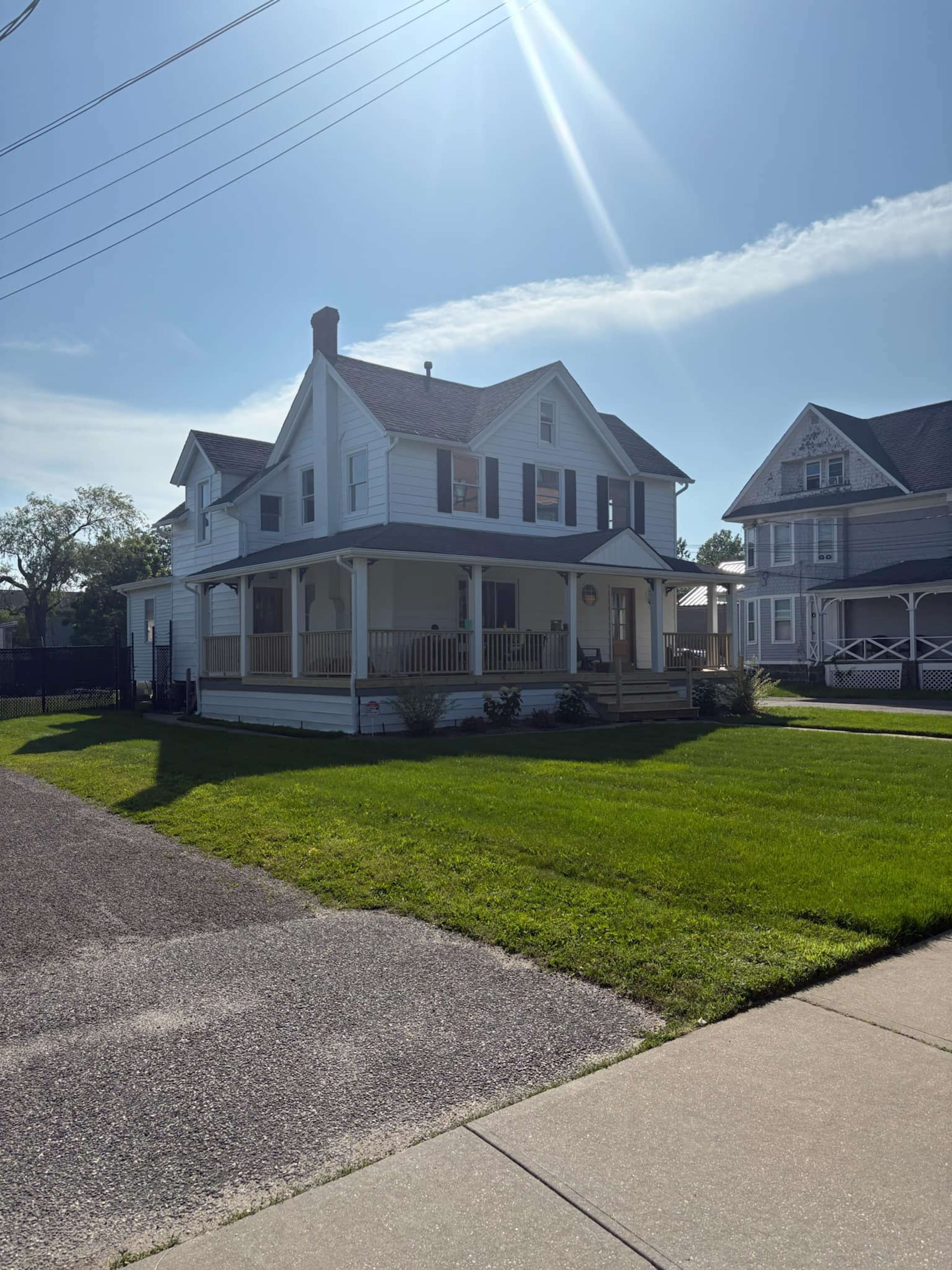 A large, two-story white house with a porch and a well-maintained lawn stands beside a concrete sidewalk on a sunny day.